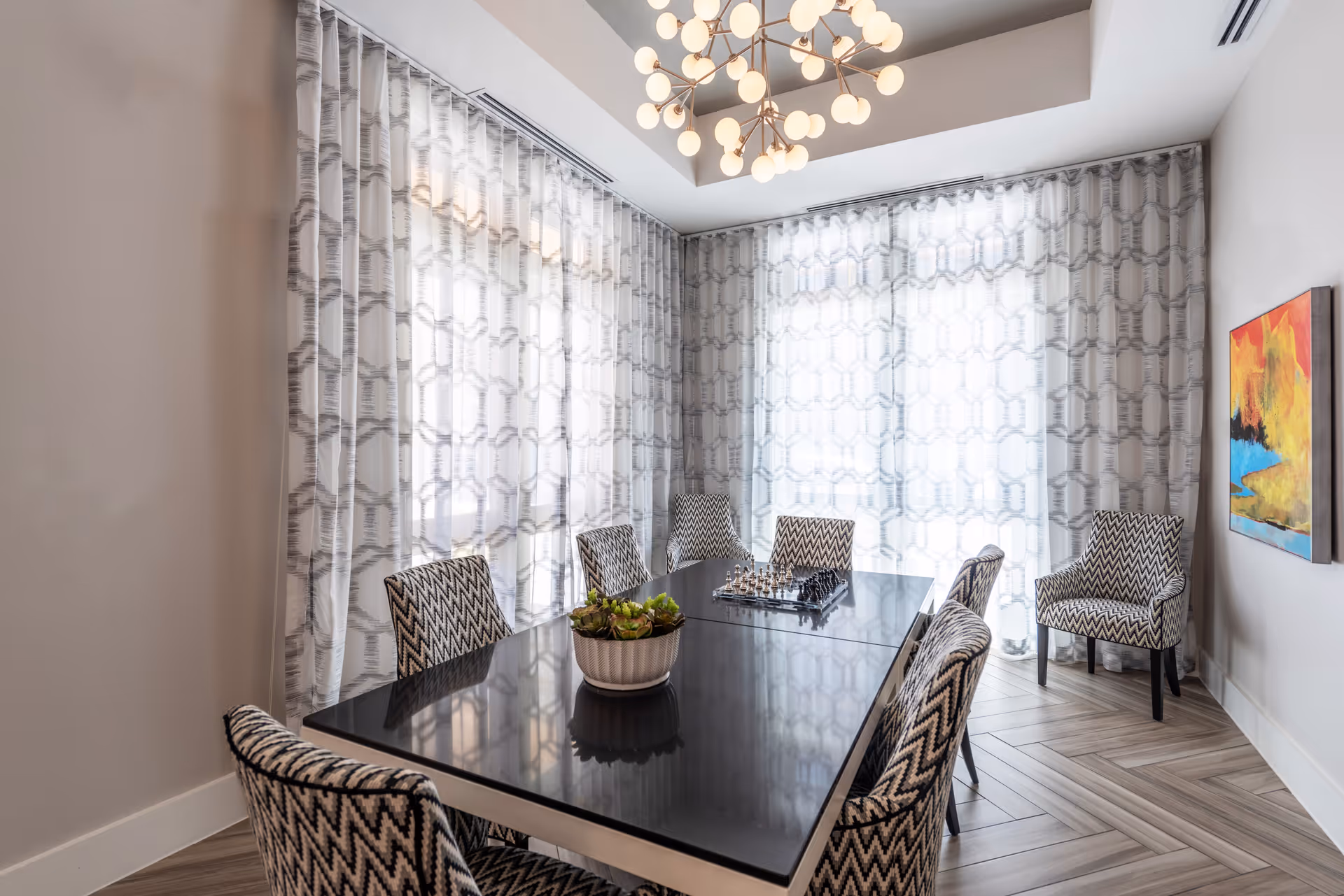A modern dining room with a black rectangular table surrounded by six patterned chairs. The room features large windows covered with sheer geometric-patterned curtains, a contemporary chandelier with multiple round bulbs, a potted plant centerpiece on the table, and a colorful abstract painting on the wall.