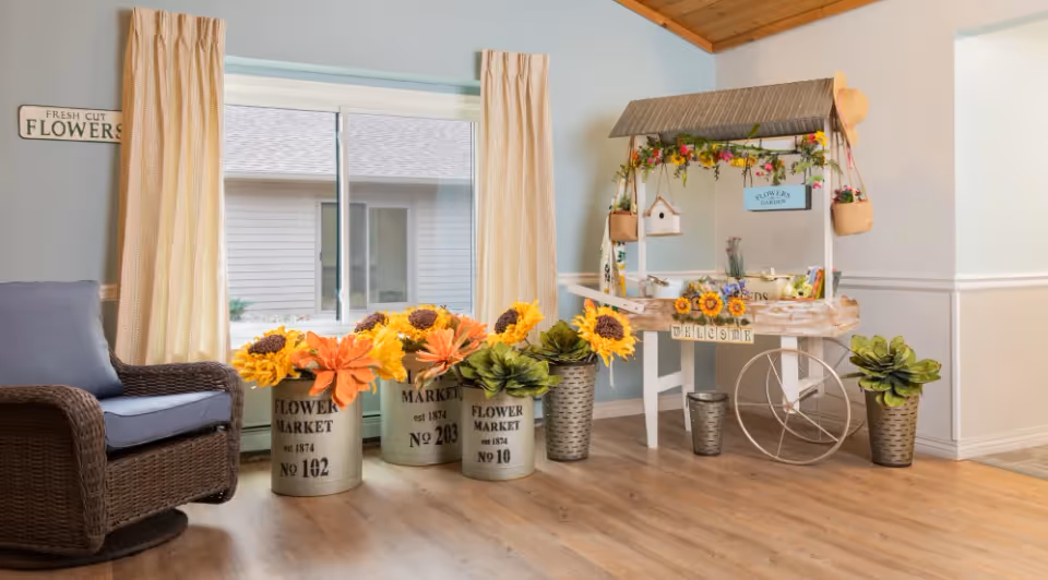 Sunlit common area featuring a decorative flower market cart, large potted sunflowers, and a wicker chair by a window.