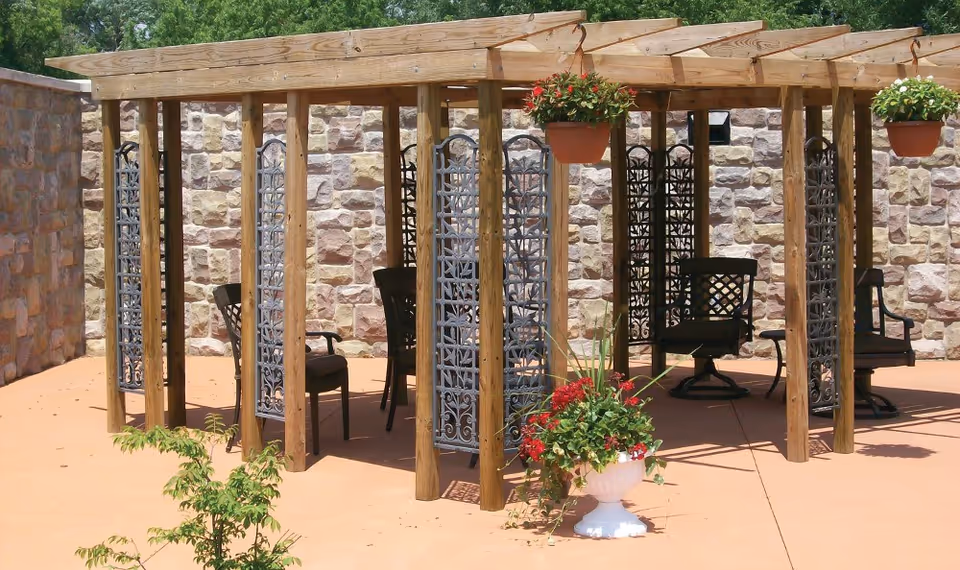Outdoor seating area with a wooden pergola structure, metal decorative panels, black chairs, and hanging flower pots with red and white flowers. The area is paved with a stone wall in the background.