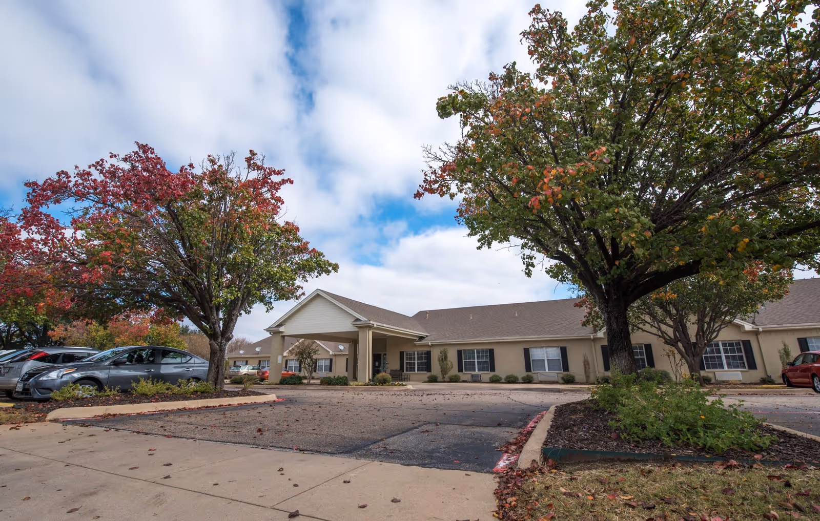 Exterior view of Ashwood Court senior living facility showing a single-story building with a covered entrance, surrounded by trees with autumn foliage and a parking area with several cars.