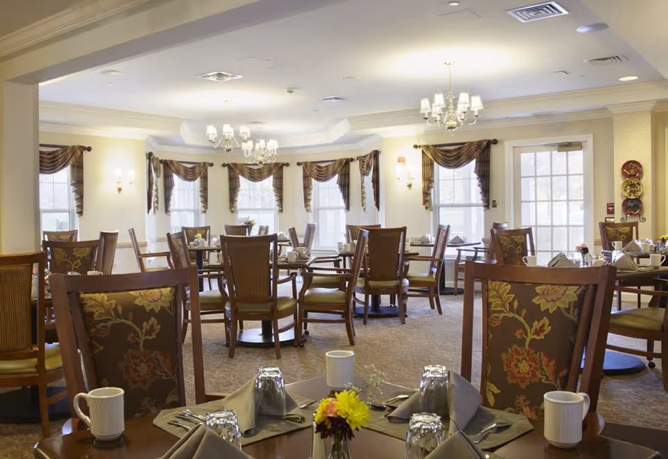 Bright dining room with multiple set tables and wooden chairs, chandeliers, and windows with drapes.