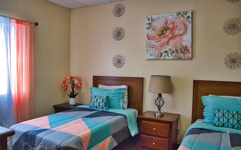A bedroom with two twin beds featuring colorful bedding in shades of teal, pink, and gray. Each bed has a decorative pillow and a wooden headboard. Between the beds is a wooden nightstand with a lamp. On the wall above the nightstand is a large floral painting and decorative wall art. A window with sheer and coral curtains is on the left side of the room.