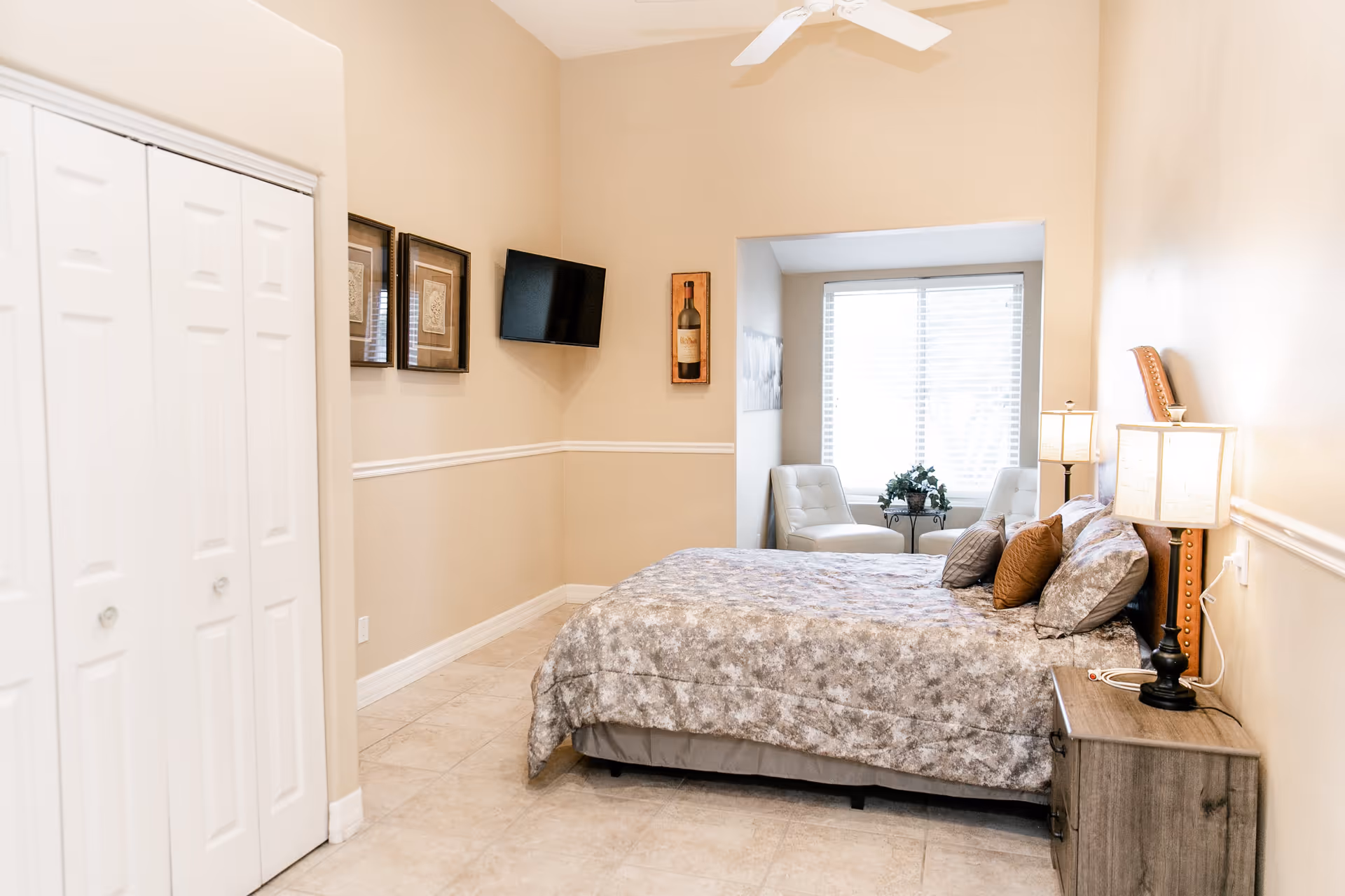 A bedroom in an assisted living facility with a bed covered in a patterned comforter and multiple pillows. There are two bedside lamps on a wooden nightstand, a wall-mounted TV, framed artwork, and a small sitting area with two white chairs and a table near a window with blinds.