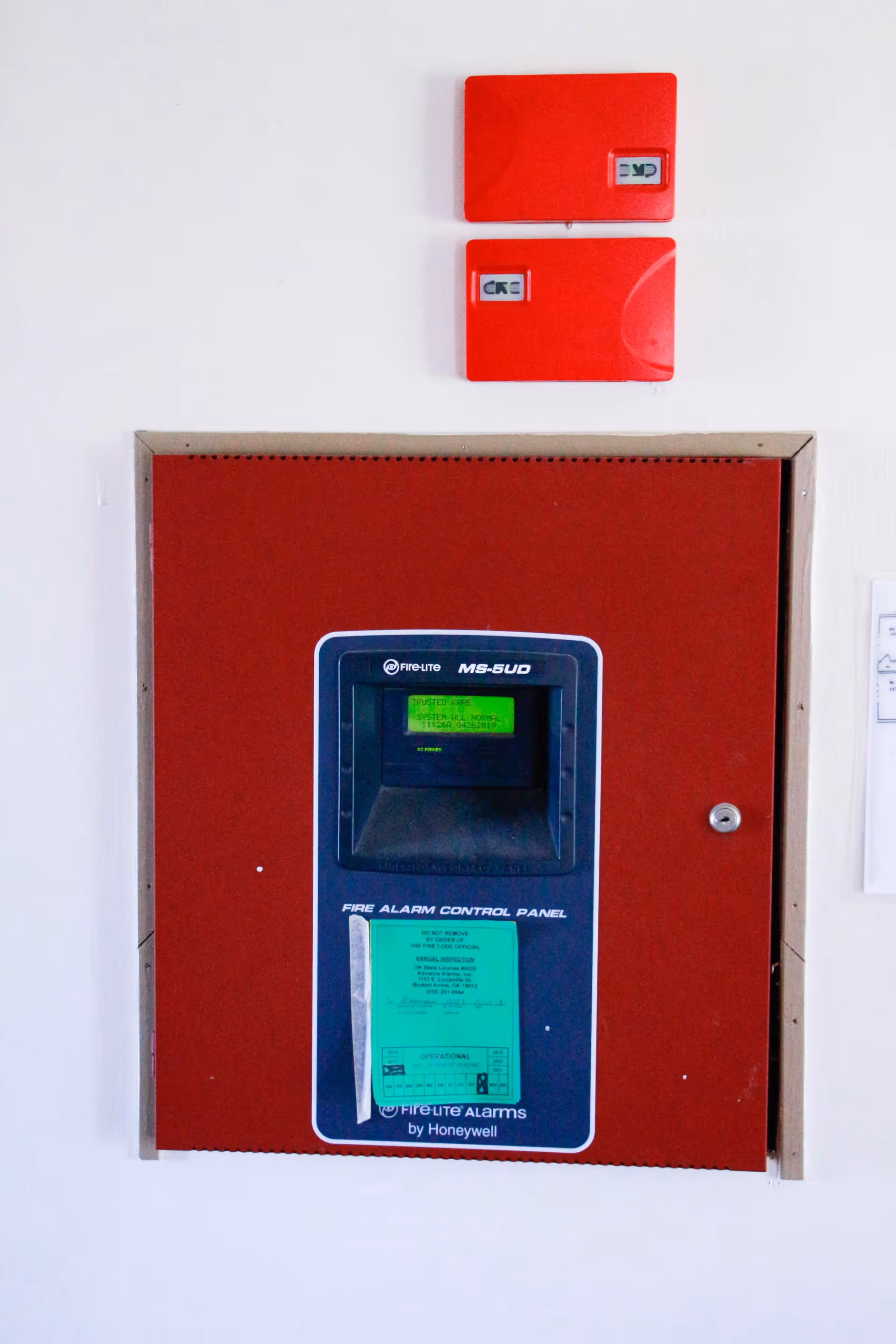 A red fire alarm control panel mounted on a white wall with two smaller red alarm devices above it. The panel has a digital display showing the text 'TRUSTED KARE SYSTEM ALL NORMAL' and a green inspection sticker attached below the display.