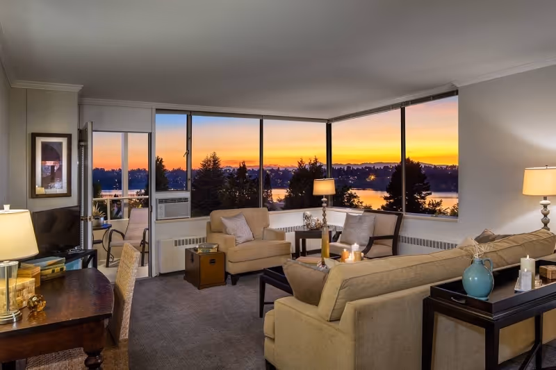 Spacious living room with beige sofas and armchairs arranged around a coffee table in front of large windows showing a lakeside sunset.