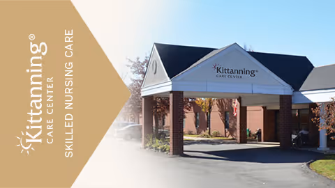 Exterior view of Kittanning Care Center showing the entrance with a covered driveway supported by brick columns, a clear blue sky, and some trees with autumn foliage.