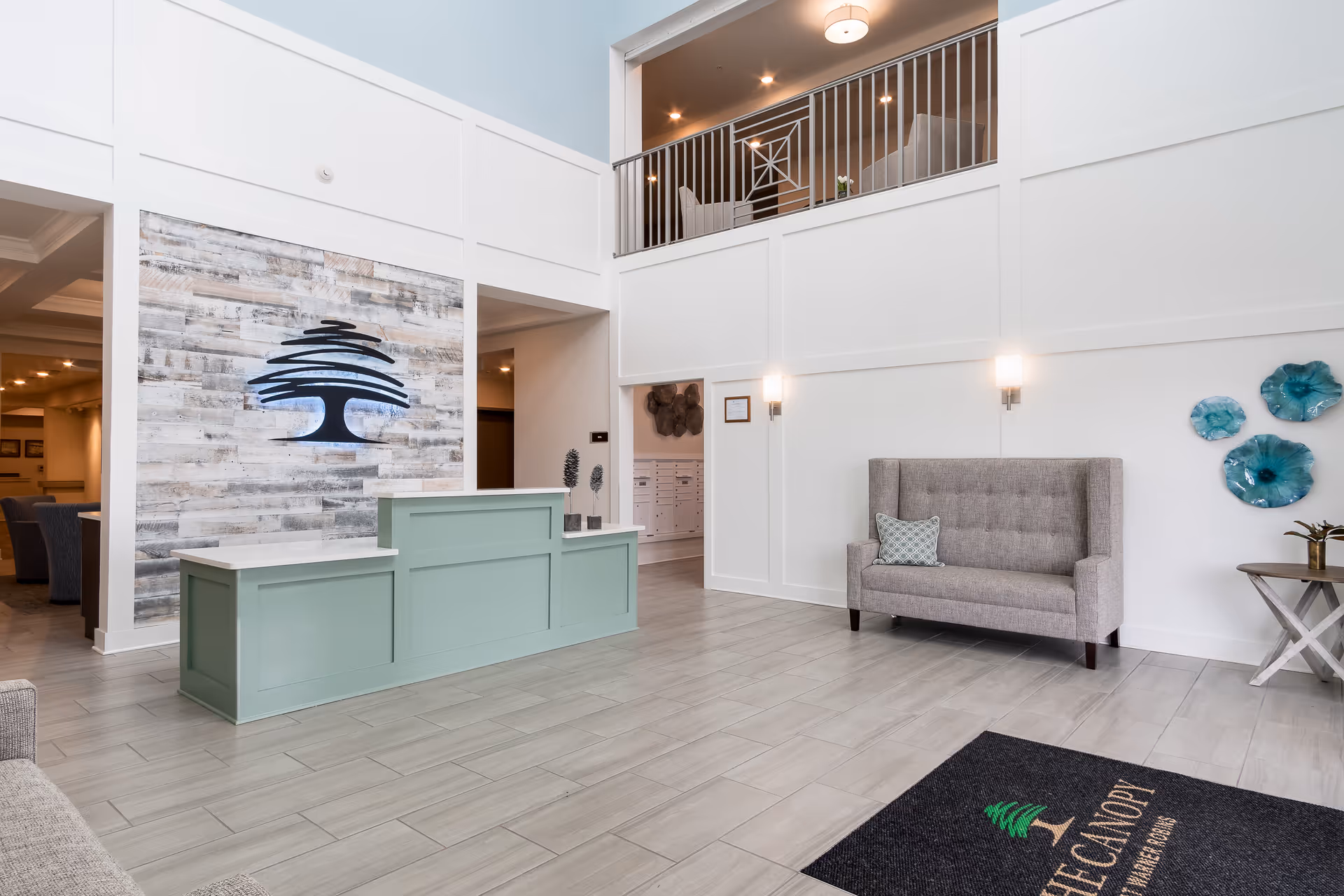A bright and spacious reception area with a light green front desk, a gray upholstered bench with a decorative pillow, and a wall adorned with blue floral art pieces. The floor is tiled in a light gray color, and there is a black mat with The Canopy logo and name near the entrance. The wall behind the desk features a large wooden panel with a tree logo, and there is a second-floor balcony with seating visible above.