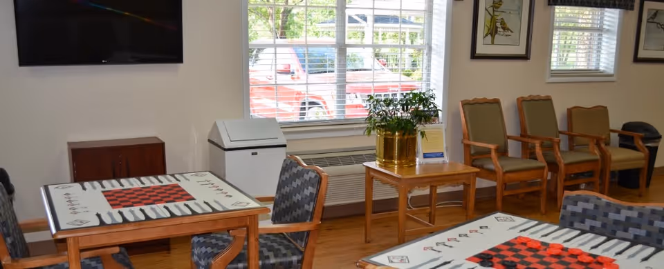 A bright room with two wooden tables featuring checkerboard and backgammon game designs, surrounded by cushioned chairs. A window with blinds lets in natural light, and a small table with a potted plant is placed near the window. Several wooden armchairs with green cushions are lined up against the wall, which is decorated with framed artwork. A flat-screen TV is mounted on the wall above a small cabinet.