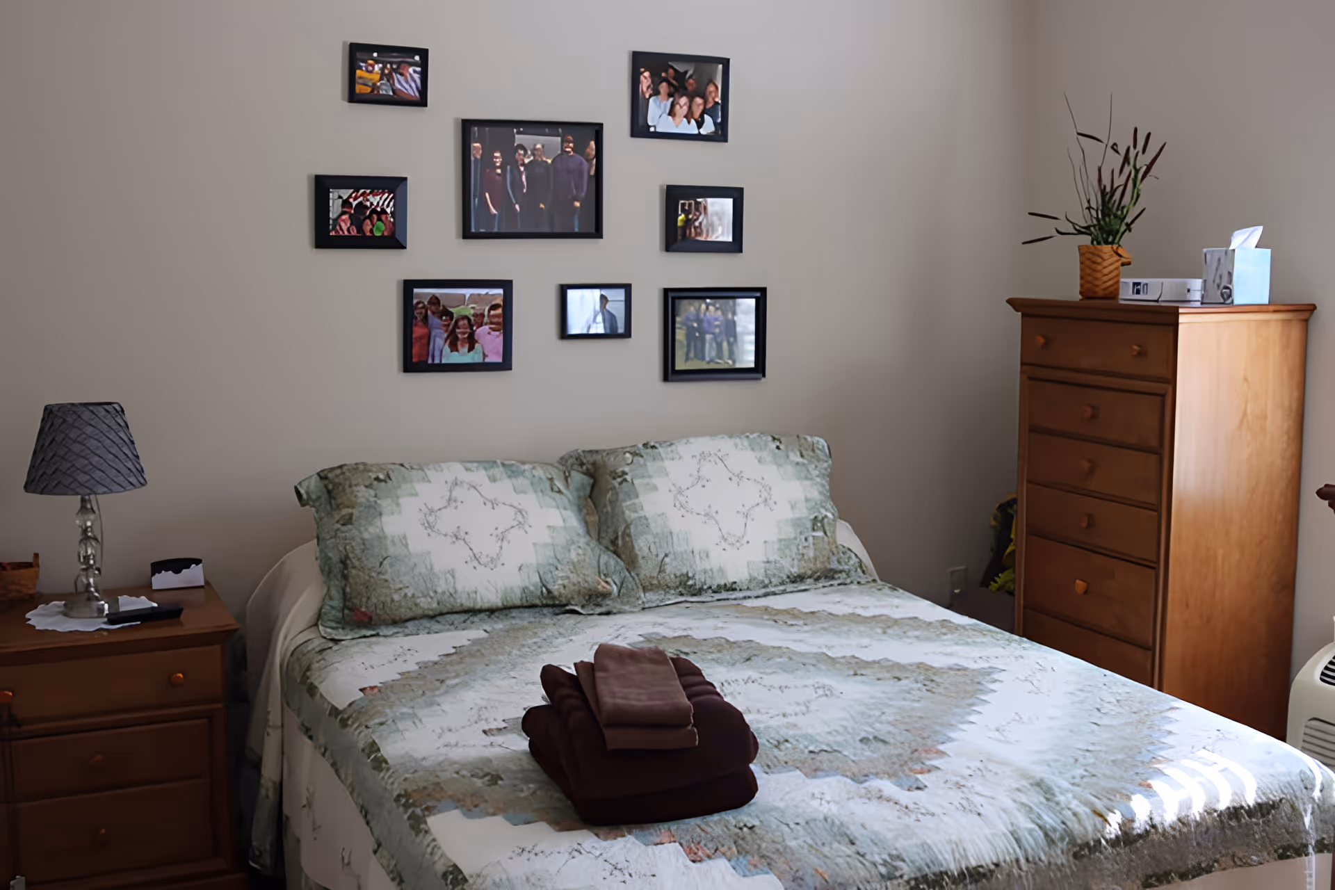 A bedroom with a neatly made bed featuring a green and white patterned quilt and two matching pillows. On the bed, there is a stack of folded towels. To the left of the bed is a wooden nightstand with a lamp and some small items. On the right side of the room, there is a tall wooden dresser with a plant and tissue box on top. The wall behind the bed has several framed photos arranged in a grid pattern.