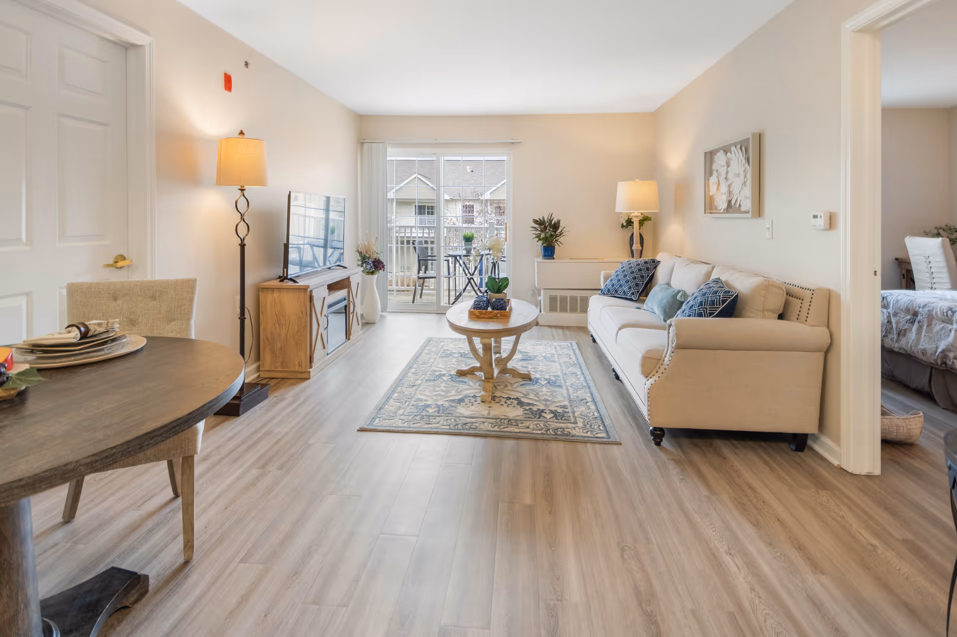 A bright living room with light wood flooring, a beige sofa with blue and green cushions, a wooden coffee table on a patterned rug, a floor lamp, a TV on a wooden stand, and a sliding glass door leading to a balcony with outdoor seating. A dining table with chairs is partially visible on the left, and a bedroom can be seen through an open doorway on the right.
