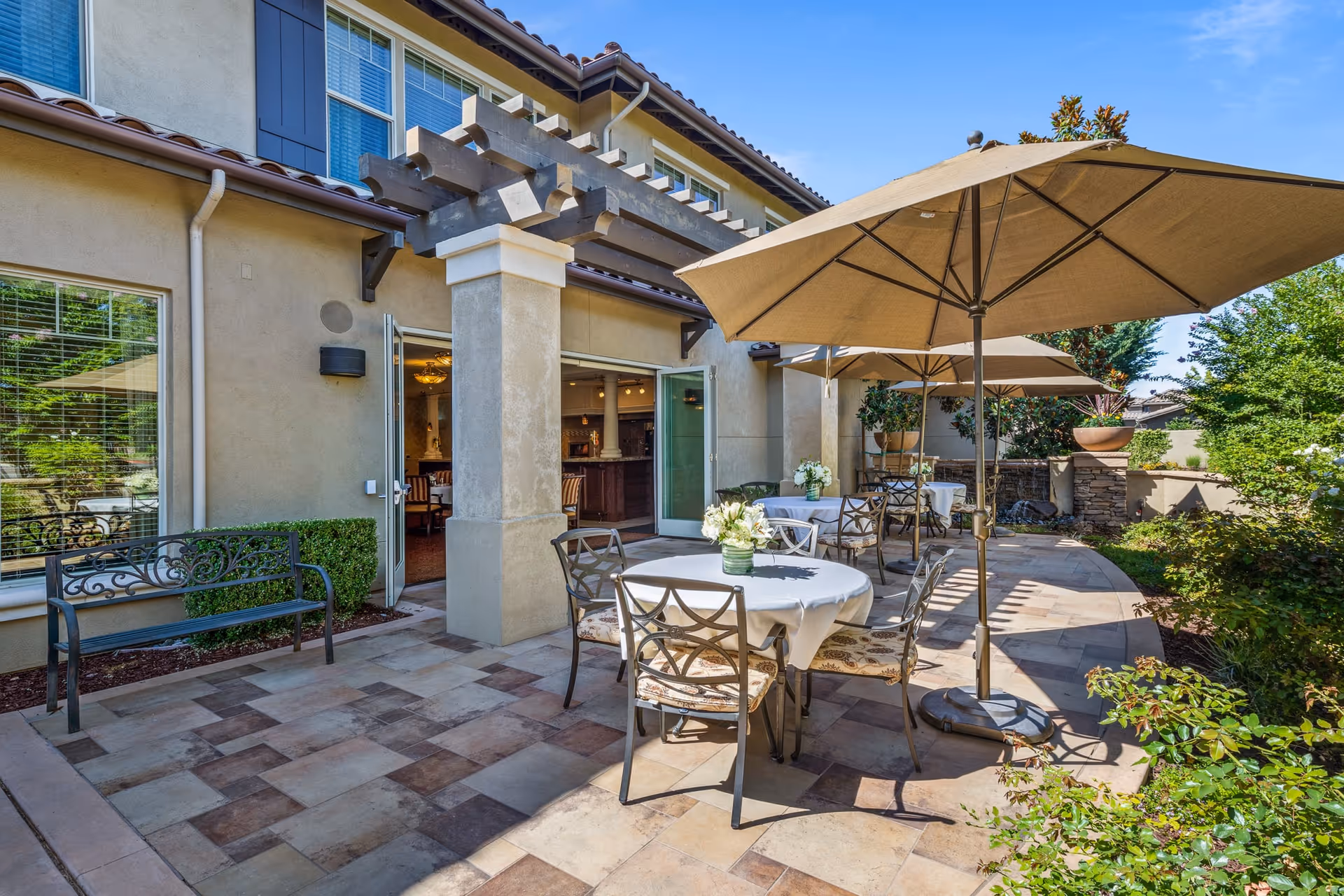 Outdoor patio area at Oakmont of Fresno with round tables covered in white tablecloths, metal chairs with cushions, large beige umbrellas providing shade, and greenery surrounding the space. The patio is adjacent to a building with large windows and an open door leading inside.