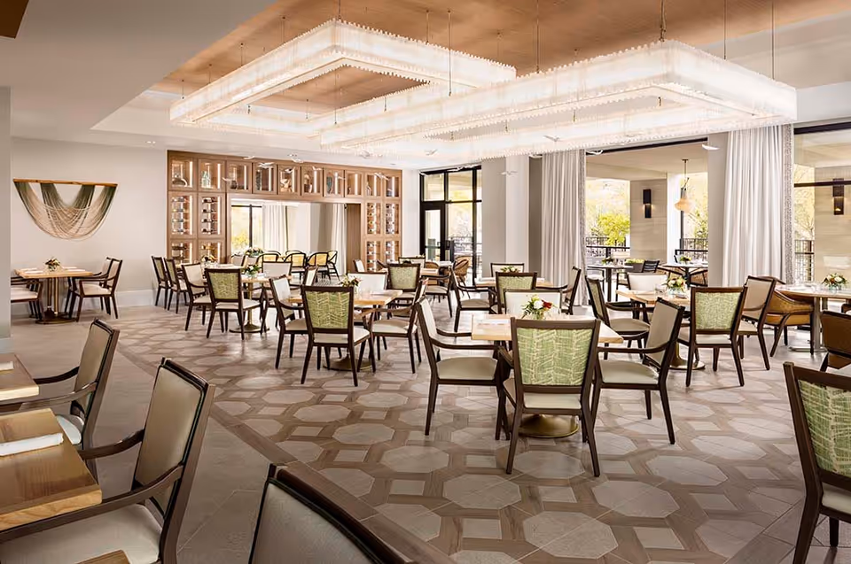 A spacious, well-lit dining room with multiple wooden tables and chairs arranged neatly. The chairs have green patterned backs and cream-colored seats. Large rectangular chandeliers hang from the ceiling, and there are large windows and glass doors letting in natural light. The room features a decorative wall with shelves and a mirror, and light-colored curtains frame the windows.
