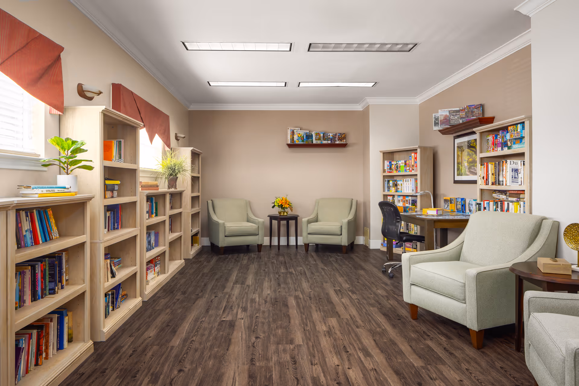 A cozy reading room with light green armchairs, wooden bookshelves filled with books and board games, a small table with a flower vase between two armchairs, and a desk with a black office chair. The room has wood flooring, beige walls, and windows with red valances letting in natural light.