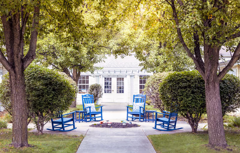 Outdoor seating area with four blue rocking chairs arranged around a small circular garden bed, surrounded by green trees and bushes, with a white building and double doors in the background.