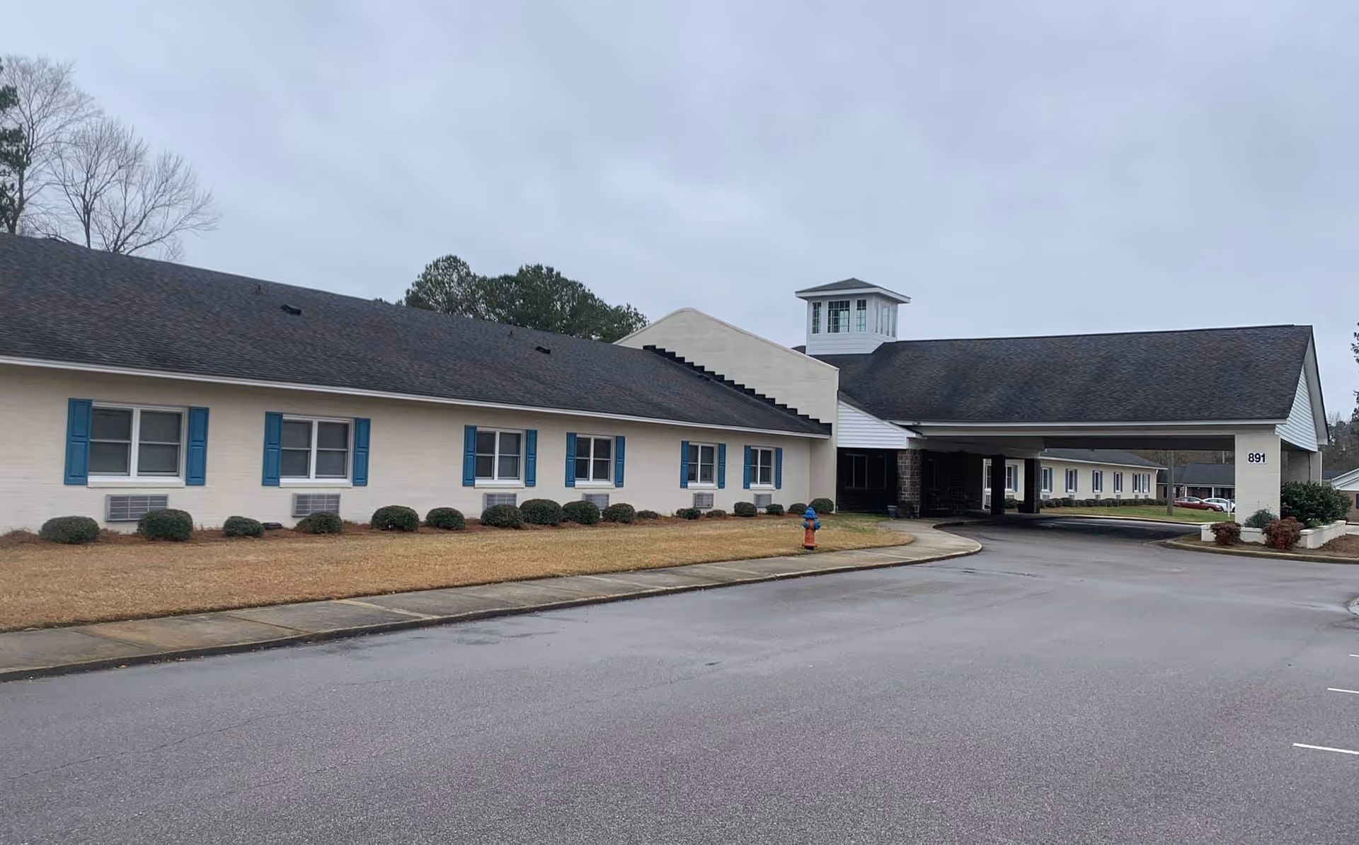Exterior view of Hunter Hill Assisted Living facility showing a single-story building with blue window shutters, a covered entrance, and a driveway. The sky is overcast and there are some trees in the background.