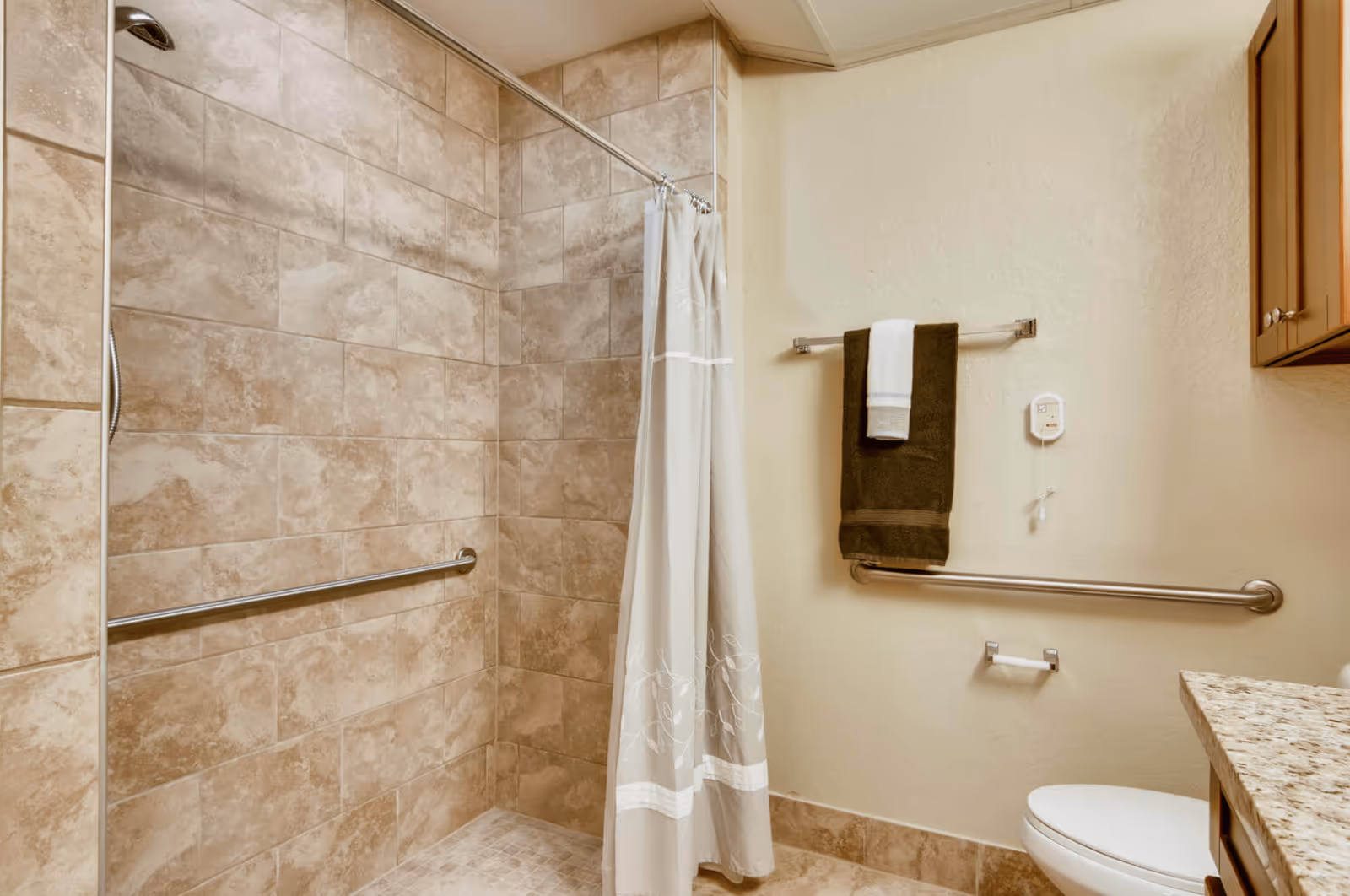 A bathroom with beige tiled walls and floor, featuring a walk-in shower with a silver grab bar and a shower curtain. There are two towel racks on the wall, one holding a dark brown towel with a white towel on top. A toilet is partially visible next to a granite countertop and wooden cabinet.