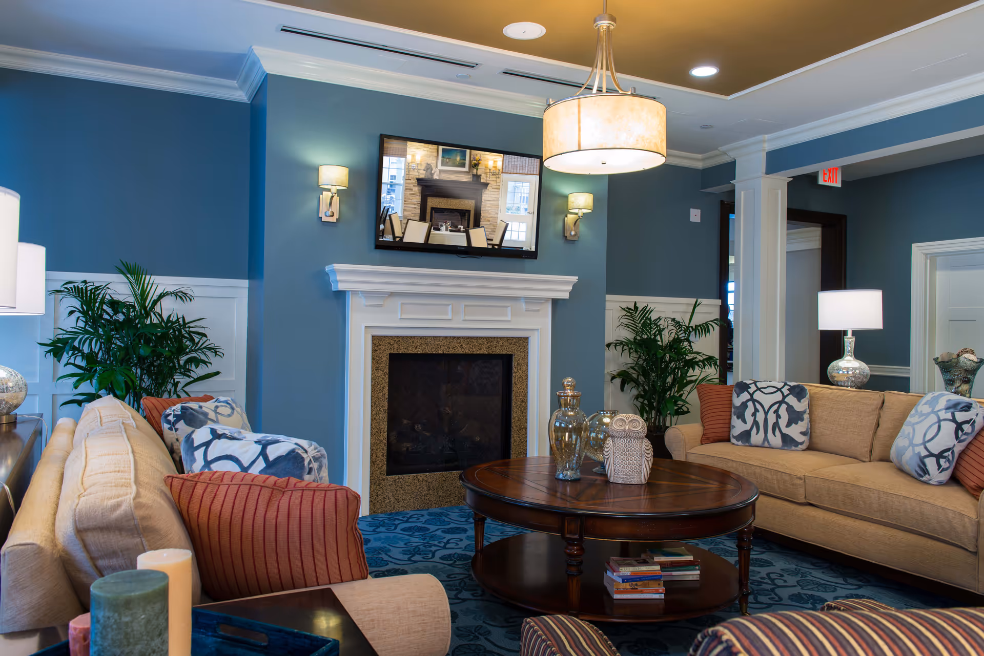 A cozy living room with beige sofas adorned with patterned and solid throw pillows, a round wooden coffee table with decorative items and books, a fireplace with a white mantel, blue walls, green plants, and warm lighting from wall sconces and a ceiling light fixture.