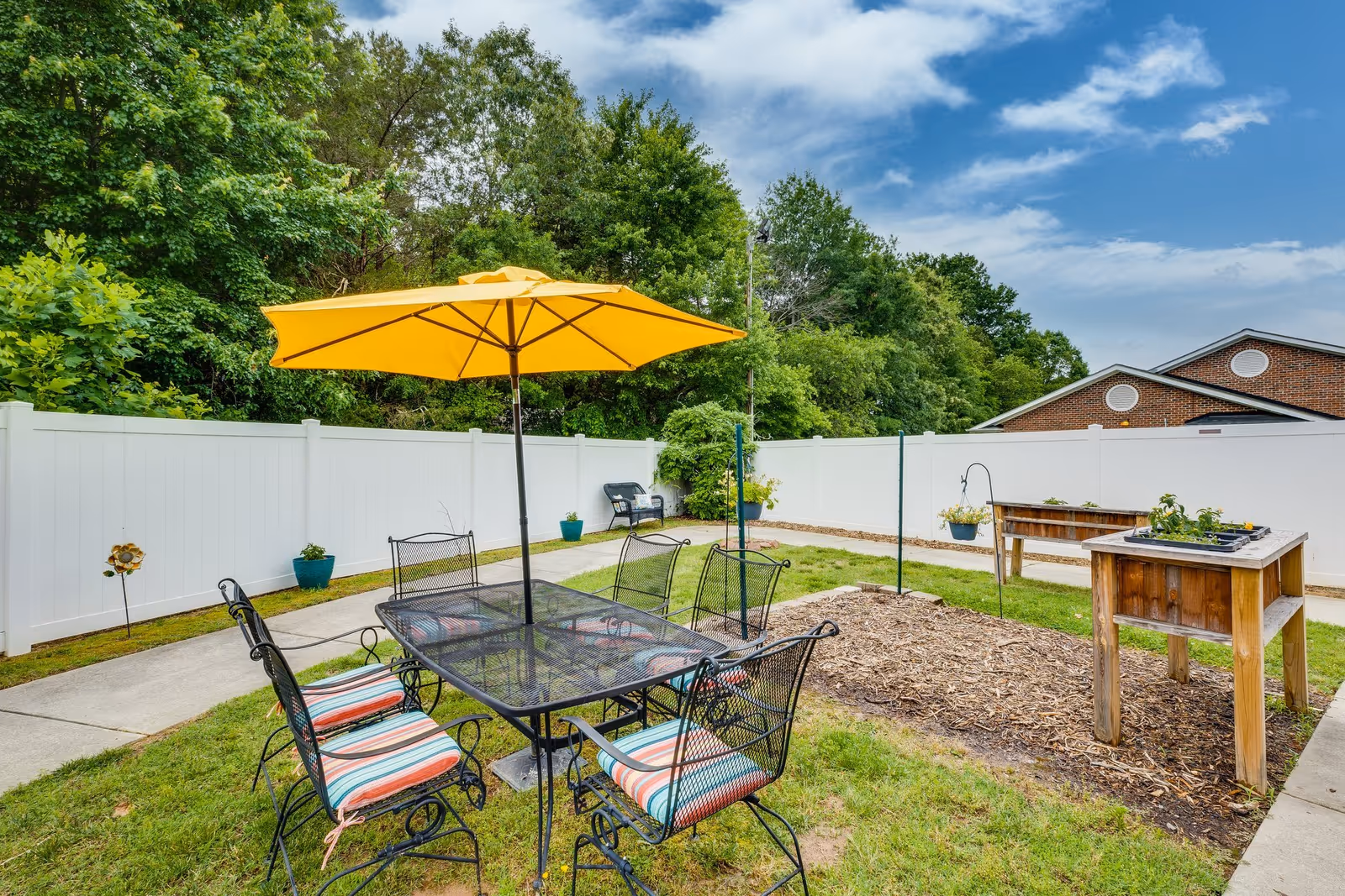 Outdoor garden area with a black metal table and six chairs with striped cushions under a large yellow umbrella. There is a white fence surrounding the area, some potted plants, a wooden raised garden bed, and a bench near the fence. Trees and a partly cloudy sky are visible in the background.