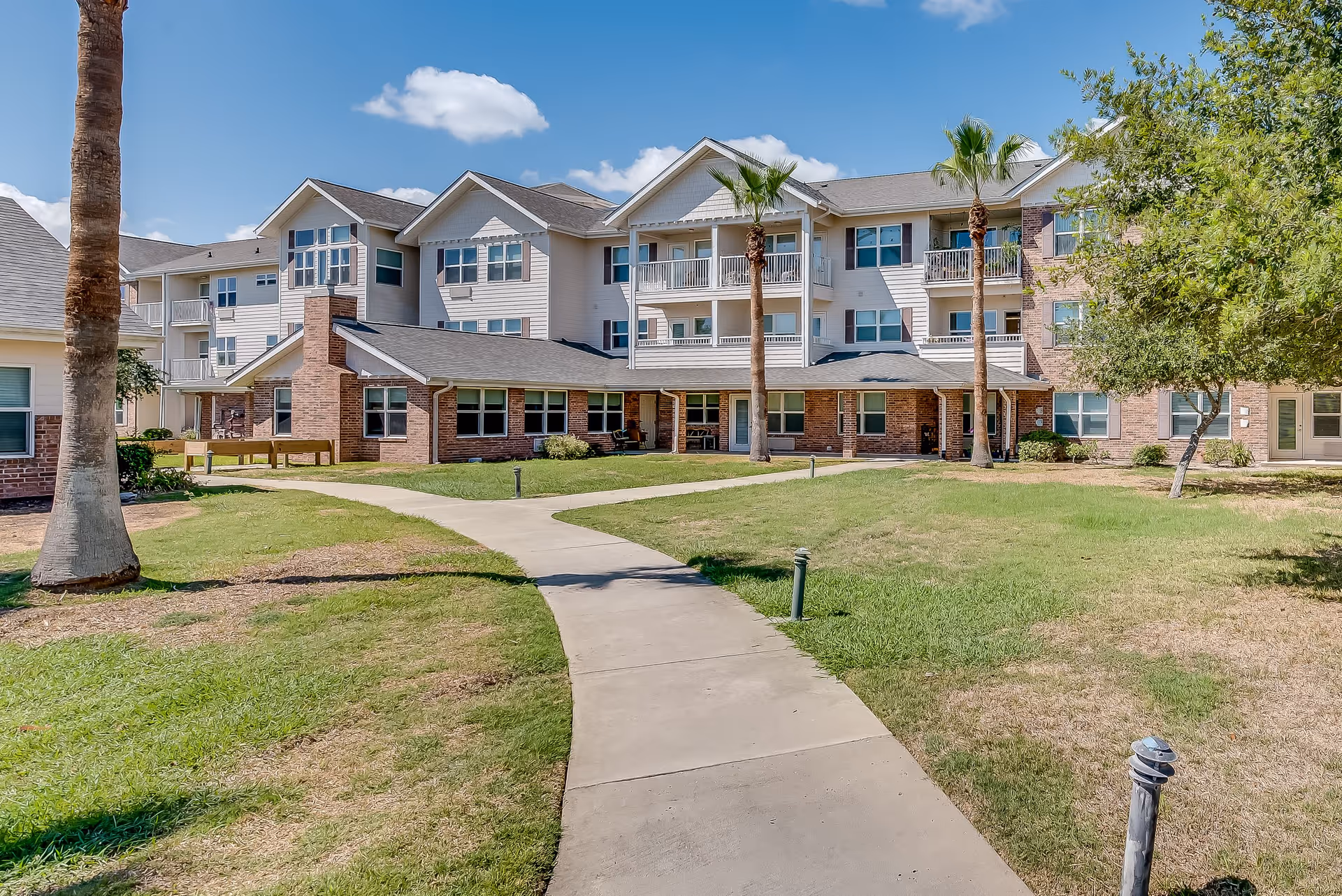 Exterior view of a multi-story senior living facility building with balconies, brick and siding facade, surrounded by a well-maintained lawn, palm trees, and a concrete walkway leading to the entrance under a blue sky with some clouds.