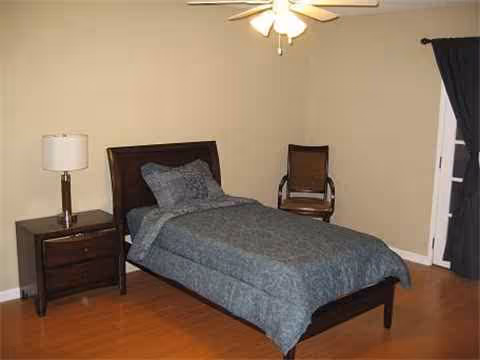 A single bedroom with a bed, nightstand, lamp, and a chair in a neutral-toned room.