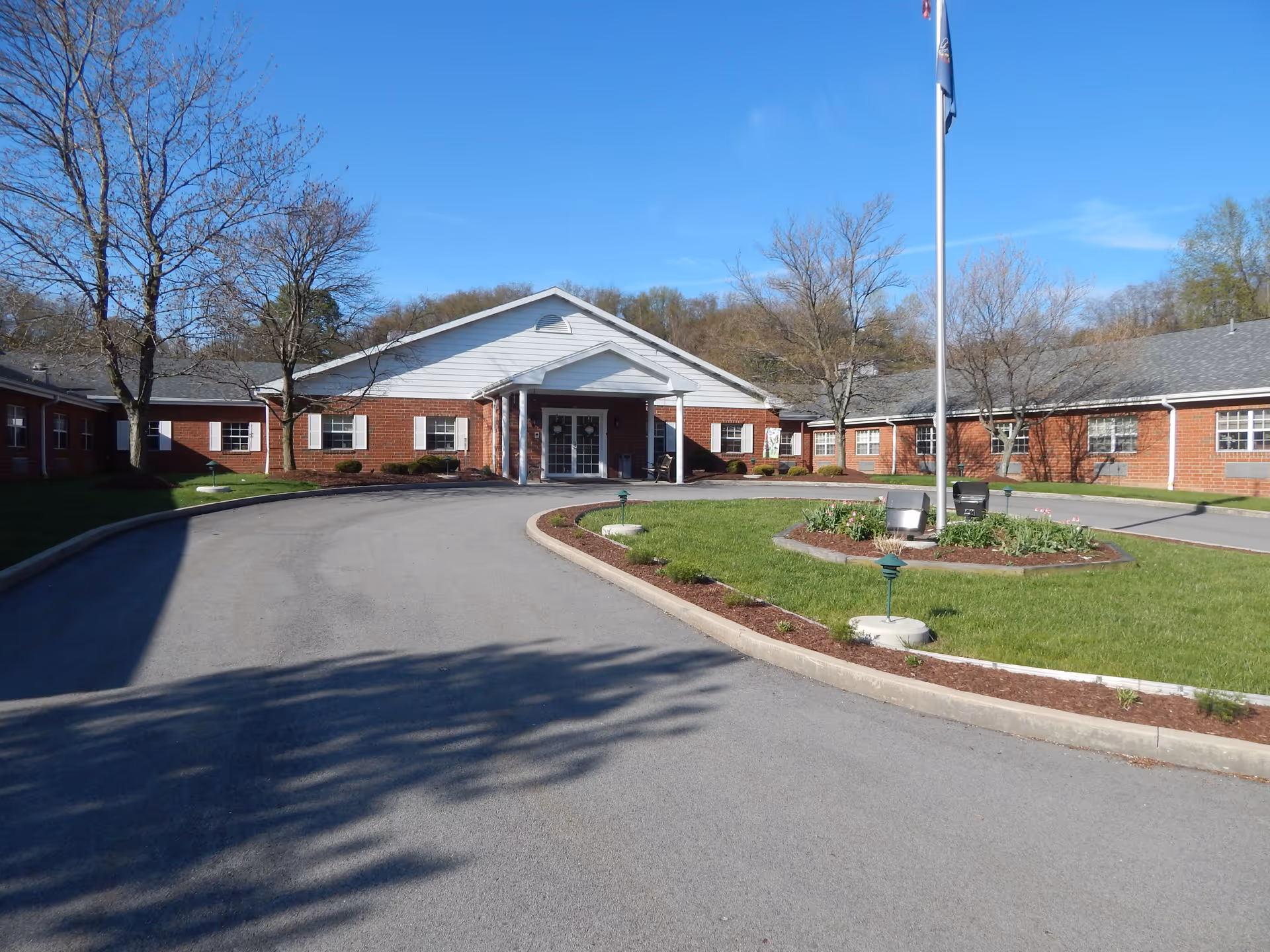Front exterior of a single-story brick senior living building with a circular driveway, flagpole, and landscaped lawn.