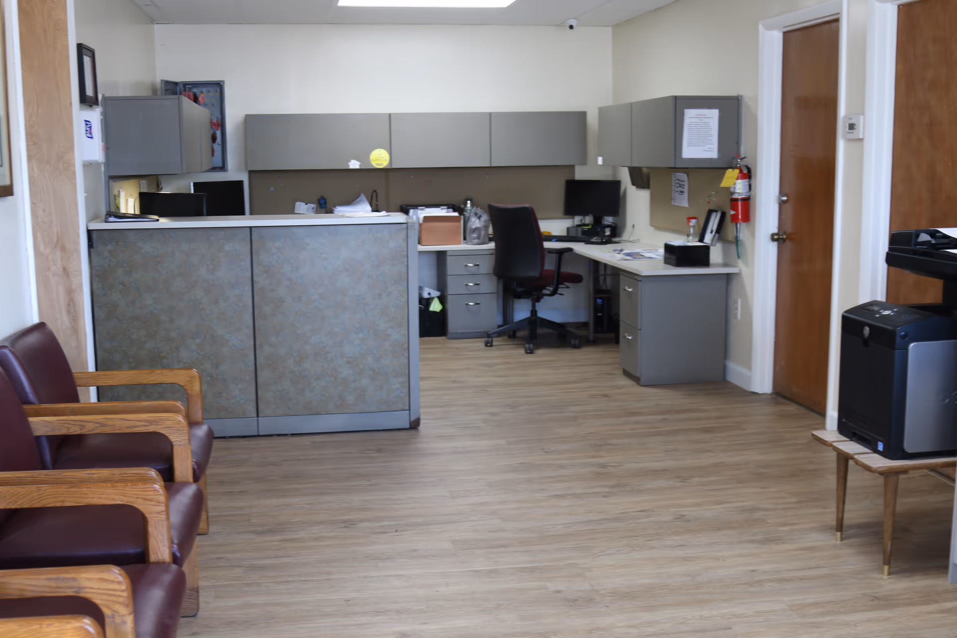 Waiting room and reception area with maroon chairs, a front desk, office workspace and a printer.