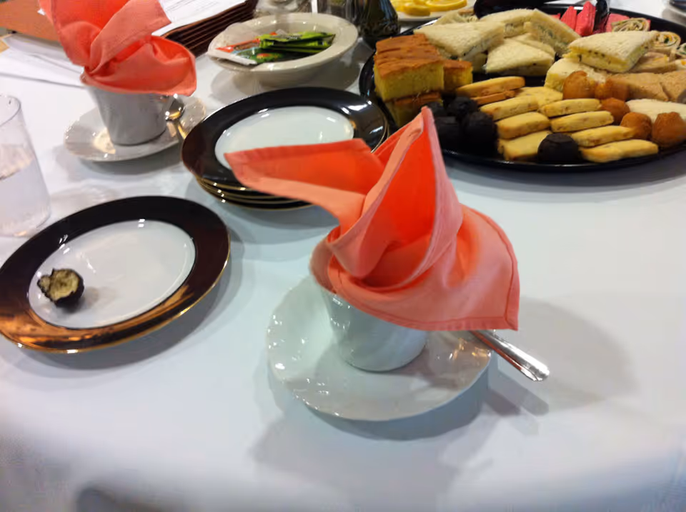 A table set for tea with cups and saucers, pink folded napkins, and a platter of sandwiches and pastries.