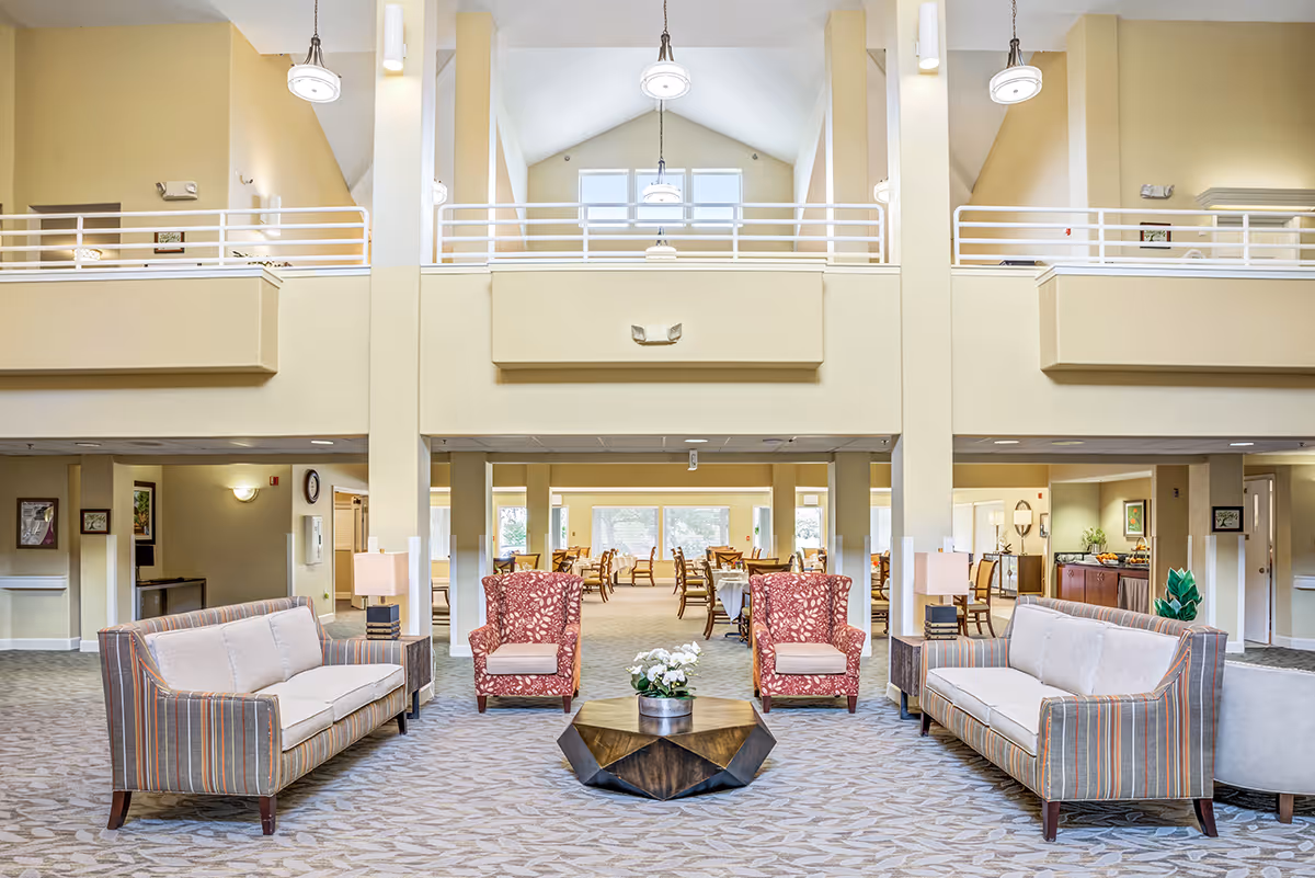 A spacious and well-lit senior living facility common area with two striped sofas and two red patterned armchairs arranged around a geometric wooden coffee table with a flower pot. In the background, there is a dining area with multiple tables and chairs, large windows letting in natural light, and a high ceiling with hanging lights.