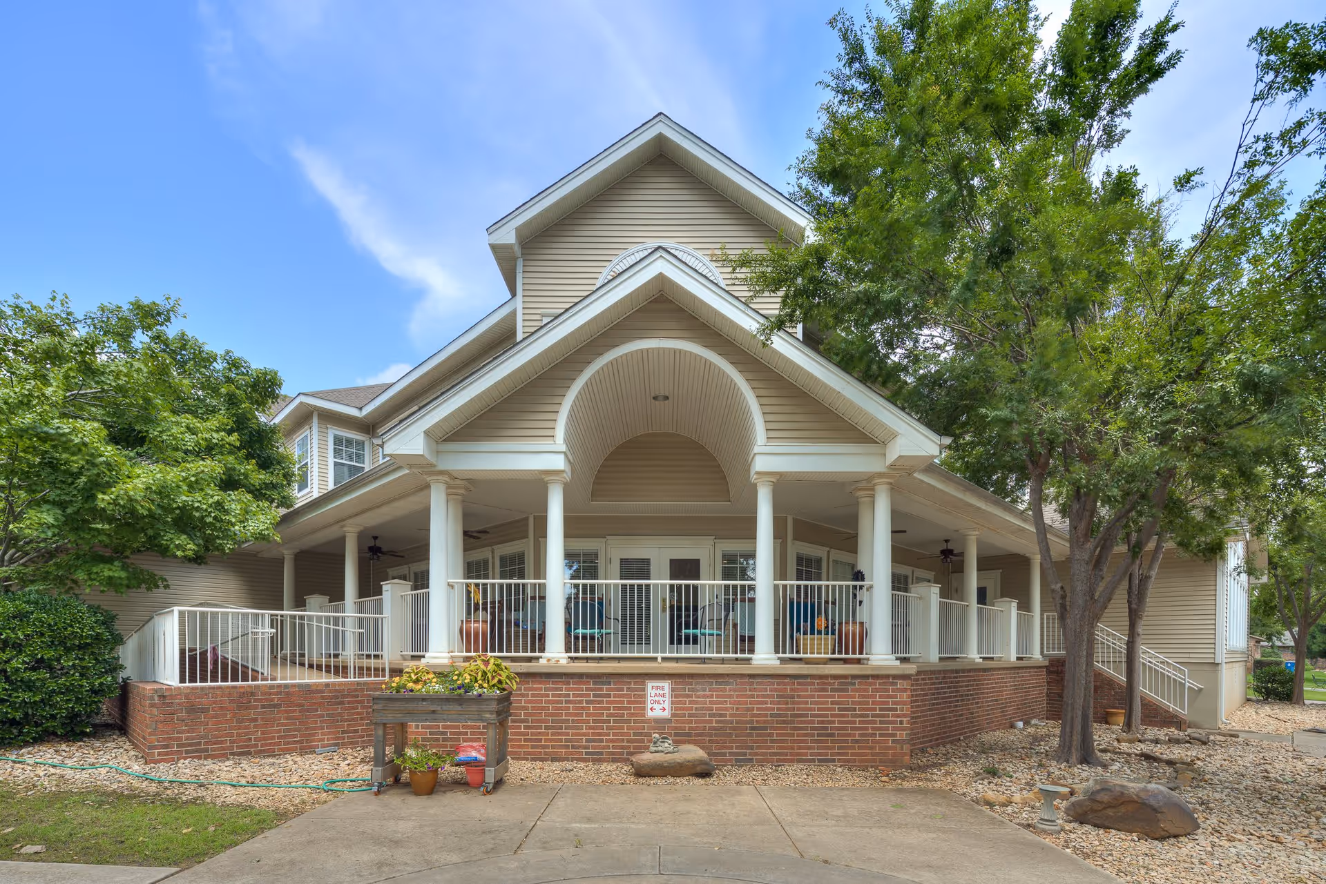 Front exterior view of a beige senior living facility building with a covered porch supported by white columns, surrounded by trees and landscaping with rocks and plants.