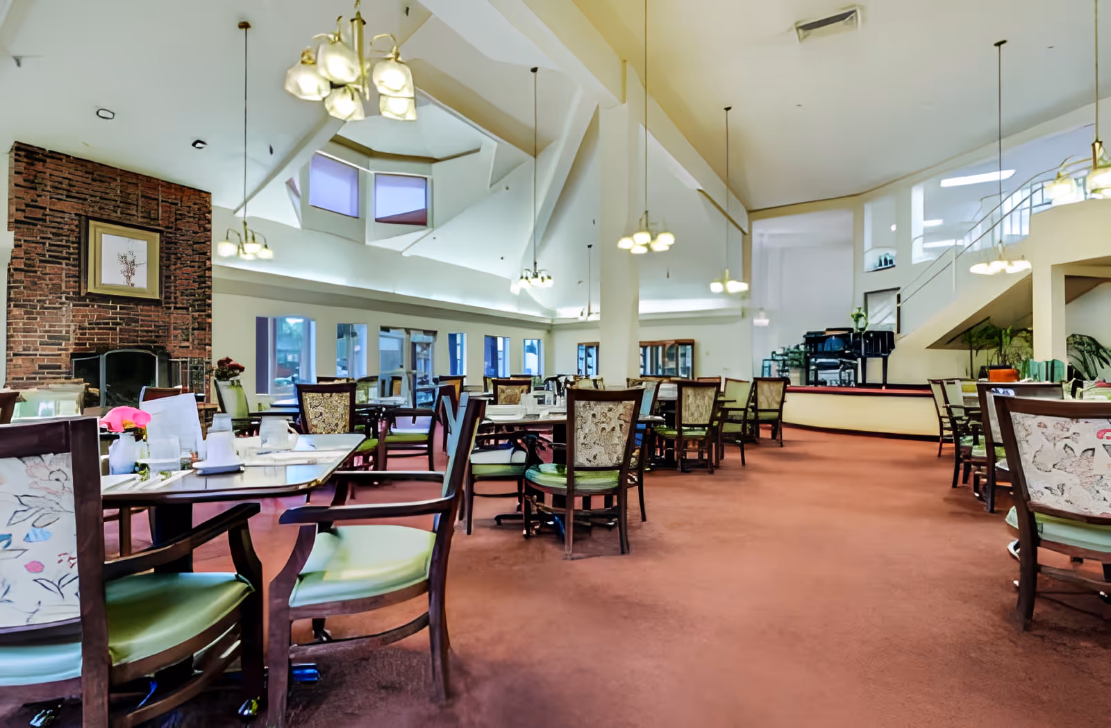 Spacious dining room with multiple tables and chairs arranged neatly on a carpeted floor. The room features a high ceiling with hanging light fixtures, a brick fireplace with a framed picture above it, large windows allowing natural light, and a grand piano near a staircase in the background.