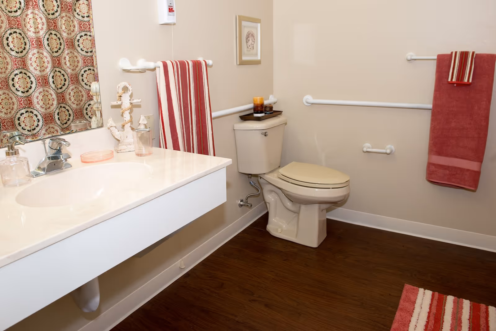 A clean bathroom with a white sink and countertop, a toilet with a beige seat, red and white striped towels hanging on white grab bars, a patterned shower curtain, and a wooden floor.
