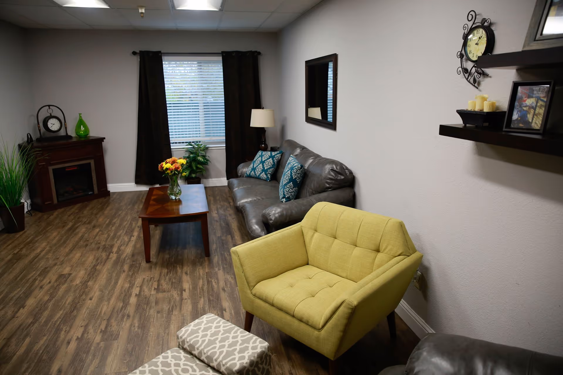 A cozy living room with wood flooring, featuring a gray leather sofa with blue patterned pillows, a yellow armchair, a wooden coffee table with a vase of flowers, a small fireplace with a clock and green vase on top, a window with dark curtains, a floor lamp, a wall mirror, and two wall shelves holding a clock, candles, and a framed picture.