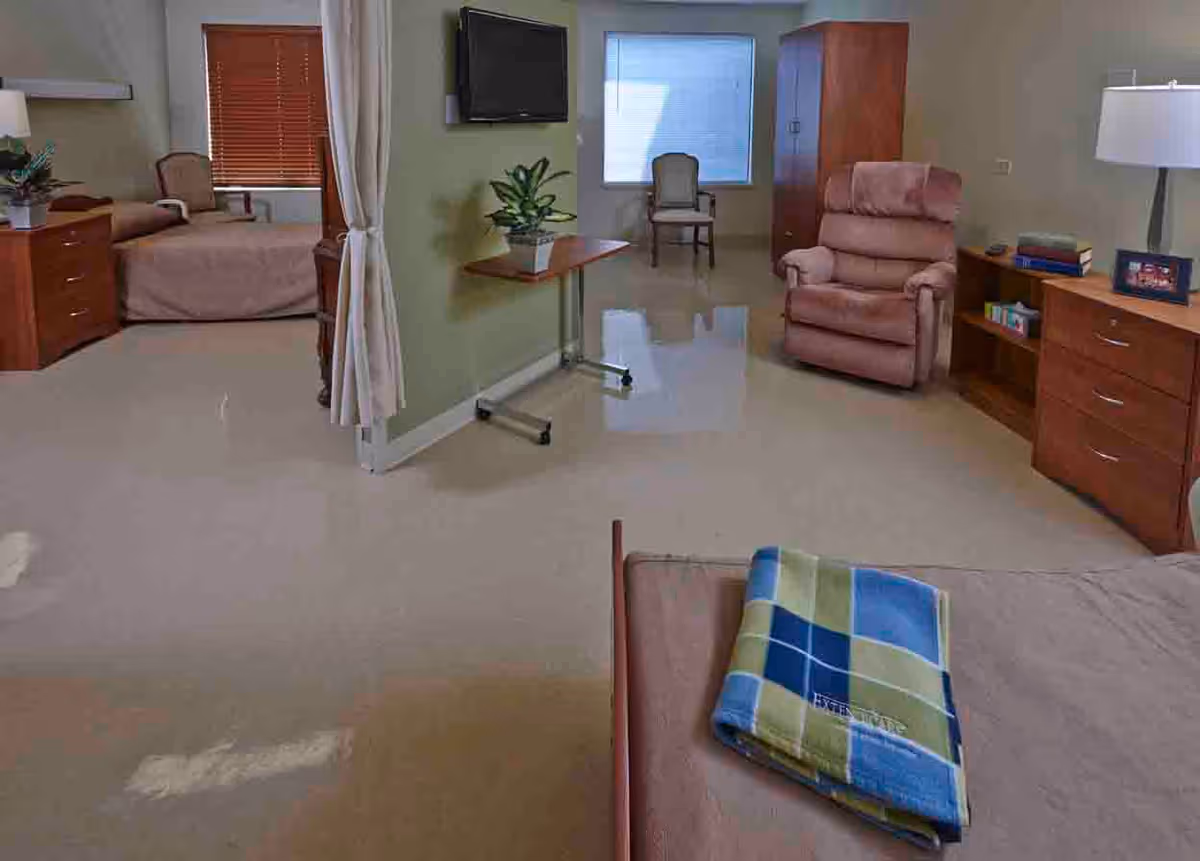 A spacious room in a senior living facility with two beds separated by a curtain. The room features wooden furniture including dressers and chairs, a recliner, a small table with a potted plant, a wall-mounted TV, and a window with blinds. A folded blue, green, and beige plaid blanket is placed on one of the beds.