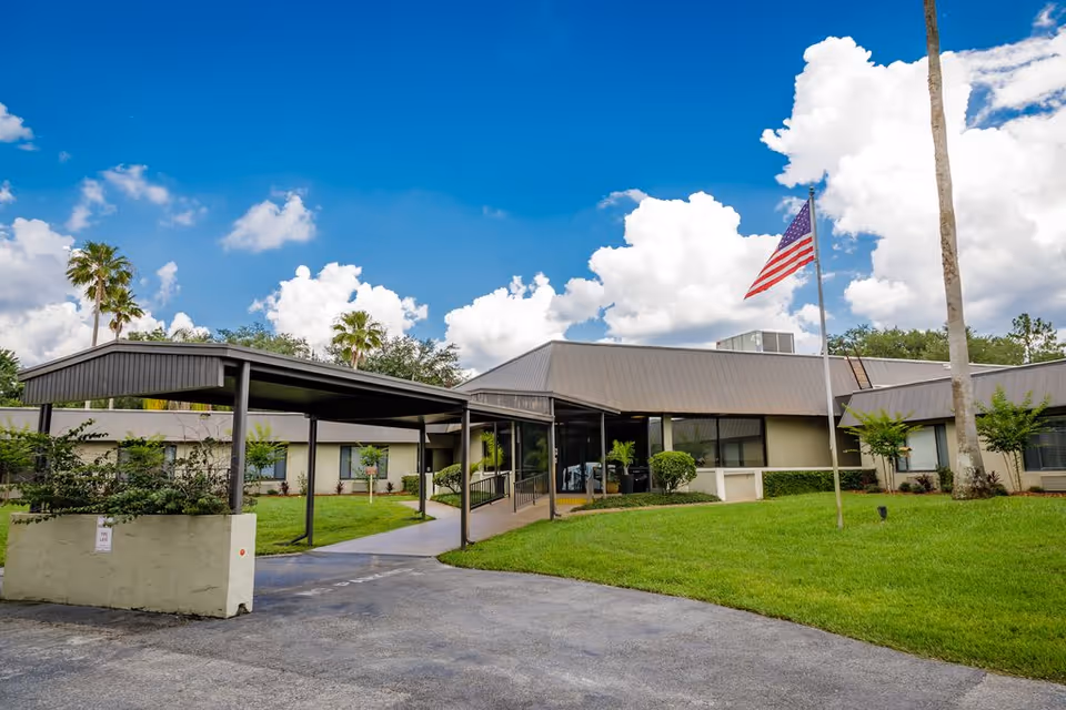 Exterior view of Solaris HealthCare Plant City facility with a covered entrance, green lawn, palm trees, and an American flag on a flagpole under a partly cloudy blue sky.