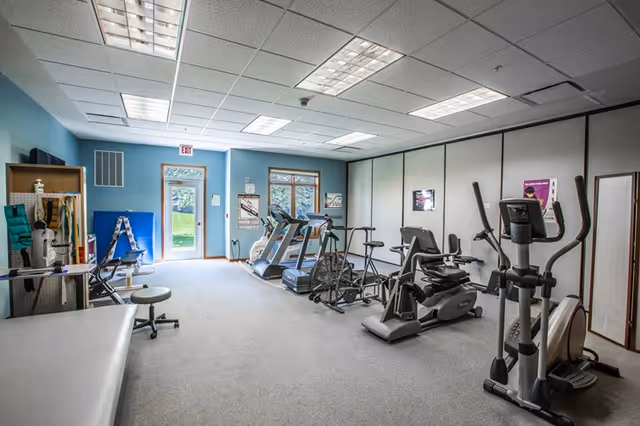 Small indoor fitness room with treadmills, ellipticals, exercise bikes, and a treatment table, with a door and windows overlooking a grassy area.