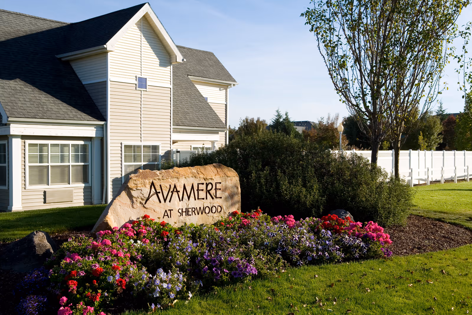 A landscaped outdoor area featuring a large rock with the inscription 'Avamere at Sherwood' surrounded by colorful flowers and greenery, with a beige building and white fence in the background under a clear blue sky.