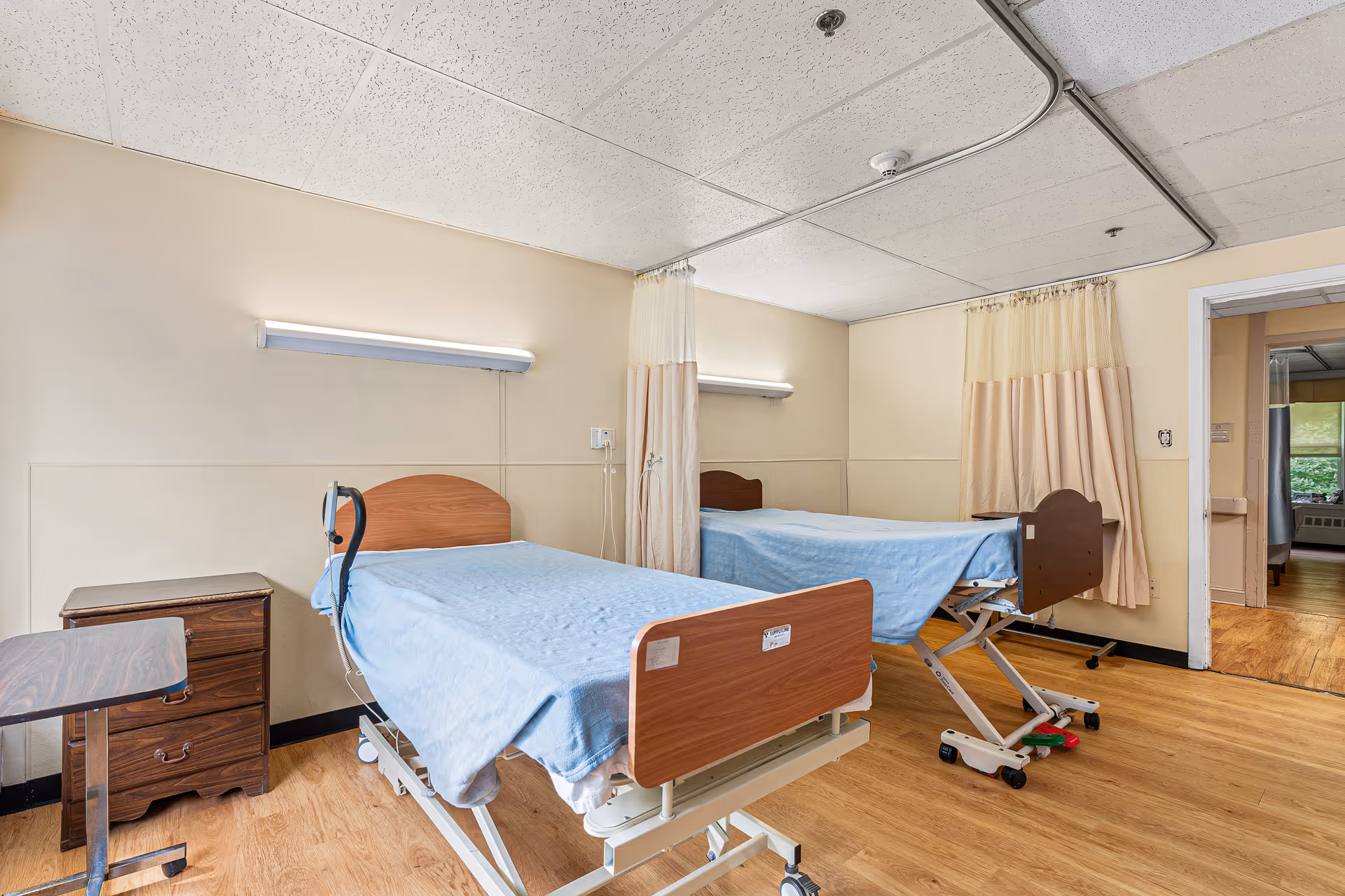 A room with two hospital-style beds covered with light blue blankets, each with a wooden headboard and footboard. There is a small wooden nightstand and an adjustable overbed table next to one bed. The room has beige walls, a drop ceiling with fluorescent lighting, and a curtain divider between the beds. The floor is wood laminate, and there is an open doorway leading to another room.