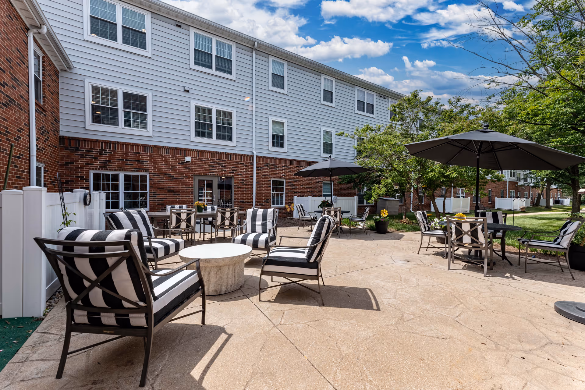 Outdoor patio area at Cedarhurst Senior Living of Tesson Heights featuring multiple seating arrangements with black and white cushioned chairs, round tables, and large umbrellas. The patio is adjacent to a brick and siding building with several windows, surrounded by green trees and a well-maintained lawn under a partly cloudy sky.
