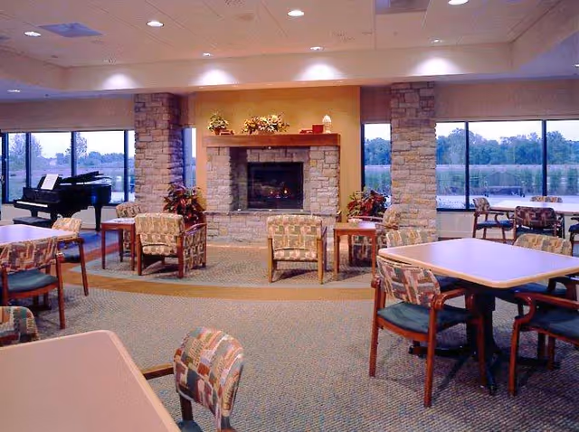 Spacious common room with tables and chairs, a stone fireplace centerpiece, large windows and a grand piano.