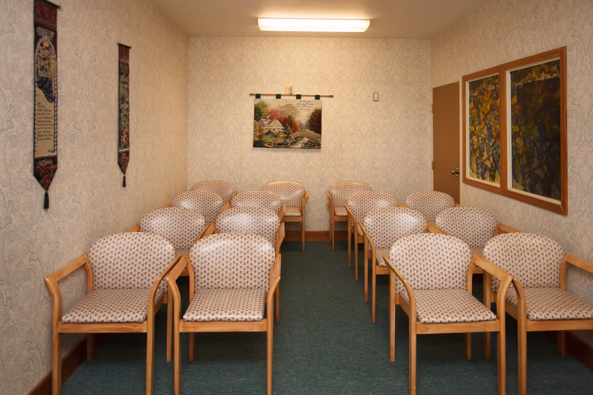 A small room with three rows of wooden chairs with patterned cushions arranged facing forward. The walls have light-colored wallpaper with a subtle pattern. On the back wall, there is a tapestry depicting a house and a bridge with trees in autumn colors. Two vertical decorative wall hangings are on the left wall, and two framed stained glass-style artworks are on the right wall next to a closed wooden door. The room is lit by a fluorescent ceiling light.