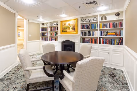 A cozy interior room with a round wooden table surrounded by four upholstered armchairs. Behind the table is a white fireplace with a framed landscape painting above it. Built-in white bookshelves filled with books and decorative items flank the fireplace. The room has beige walls with white wainscoting and a patterned carpet.