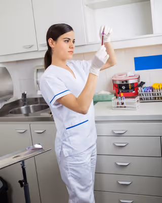 A healthcare professional wearing white scrubs and gloves is examining a test tube in a clinical setting with cabinets, a sink, and medical equipment in the background.