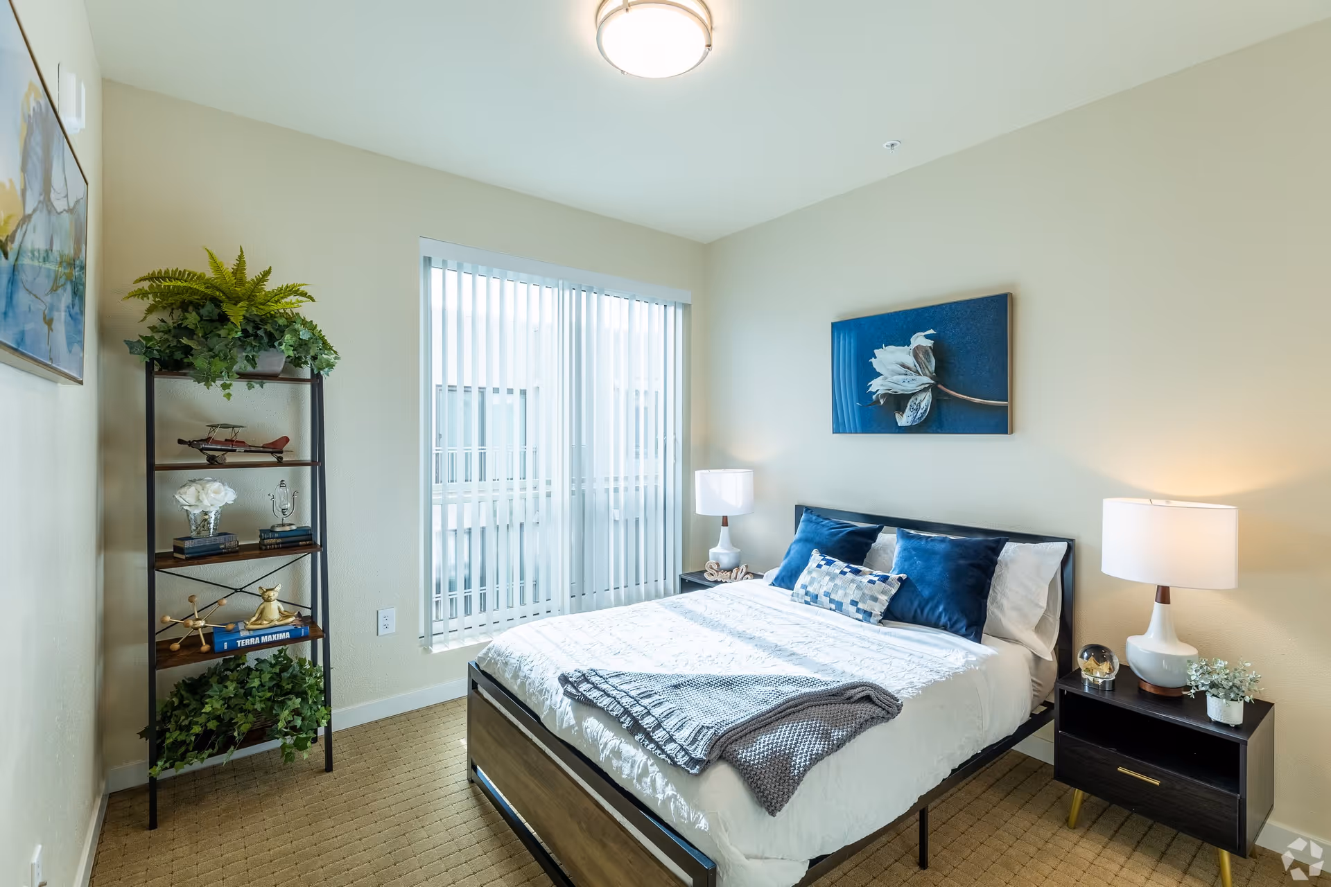 A bright bedroom with a neatly made bed featuring white bedding and blue pillows. There is a gray knitted throw blanket folded at the foot of the bed. On the right side of the bed is a dark wooden nightstand with a white lamp, a small plant, and a decorative item. On the left side of the room is a tall black metal shelf with wooden shelves holding plants, books, and decorative items. A large window with vertical blinds allows natural light to fill the room. The walls are painted light beige, and there is a framed floral artwork hanging above the bed.