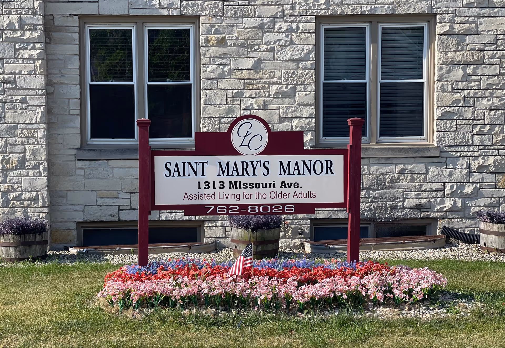 A sign for Saint Mary's Manor, an assisted living facility for older adults, located at 1313 Missouri Ave. The sign is mounted on red posts and surrounded by a flower bed with red, pink, and purple flowers and a small American flag. The background shows a stone building with two windows.
