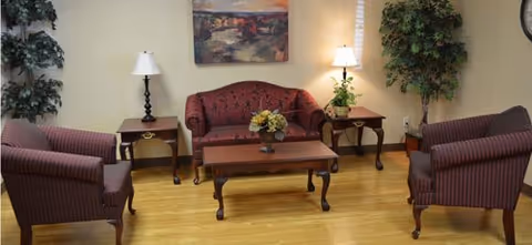 A cozy living room area with a red patterned loveseat, two matching armchairs, a wooden coffee table with a flower arrangement, two side tables each with a lamp, a large potted plant in the corner, and a framed landscape painting on the wall.