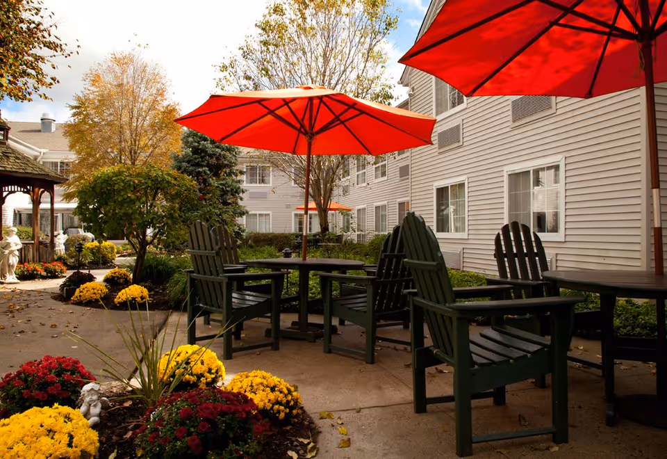 Outdoor patio area at a senior living facility with green wooden chairs and tables under large red umbrellas. The area is surrounded by colorful flower beds with yellow and red flowers, trees with autumn foliage, and a beige building with multiple windows in the background.