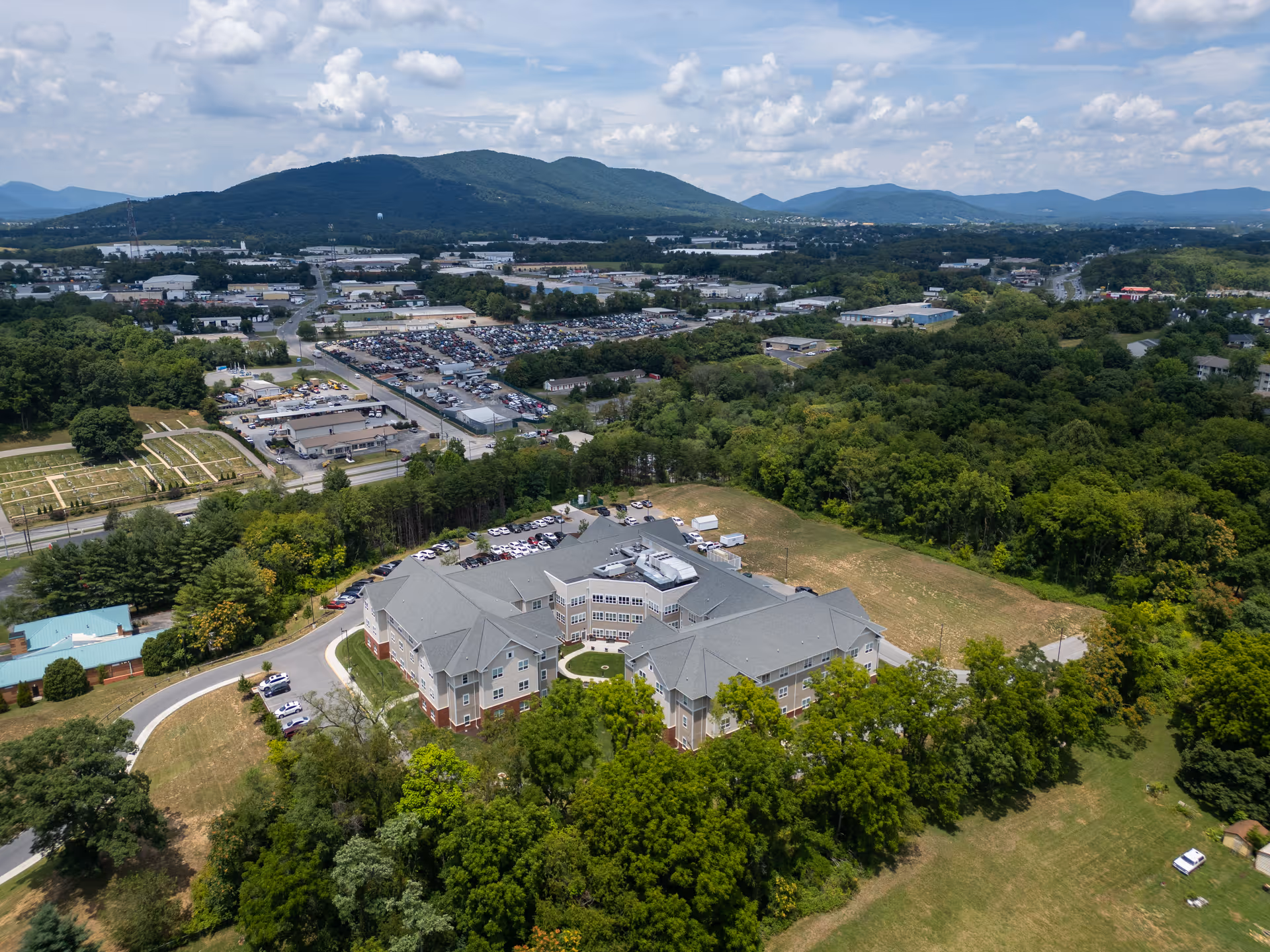 Aerial view of a large building complex surrounded by trees and open grassy areas, with a parking lot filled with cars. In the background, there are more buildings, roads, and mountains under a partly cloudy sky.