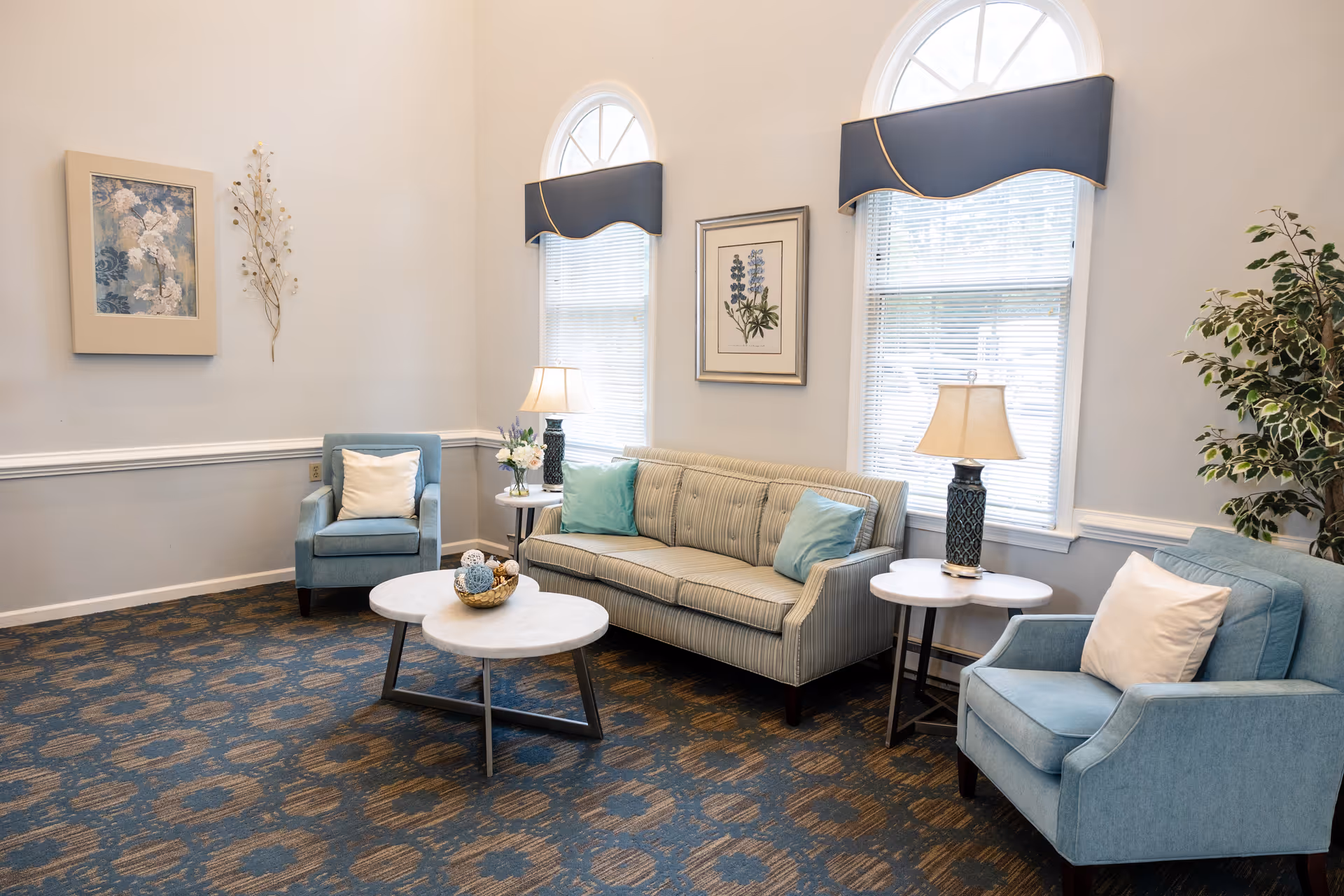 A cozy living room with two blue armchairs and a striped sofa with light blue pillows. There are two round white coffee tables in front of the sofa, each with decorative items. The room has two tall arched windows with blue valances and white blinds, two table lamps on side tables, framed floral artwork on the walls, and a potted plant in the corner. The carpet has a blue and beige pattern.