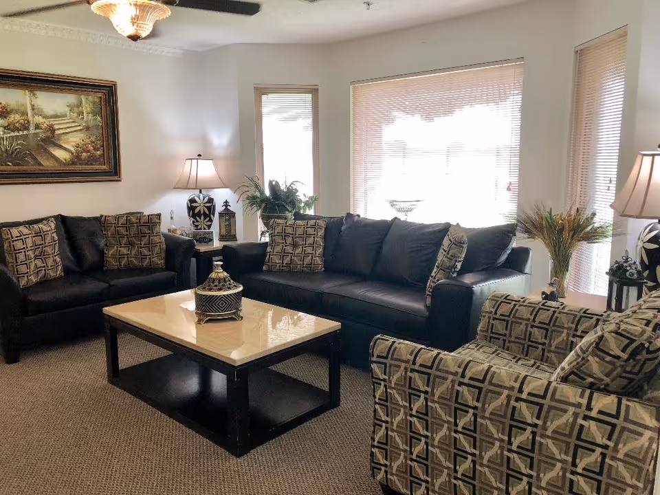 A well-lit living room with two black leather sofas and one patterned armchair arranged around a rectangular coffee table with a decorative item on top. The room has beige carpet, two table lamps with patterned bases, a large window with blinds, and a framed painting on the wall.