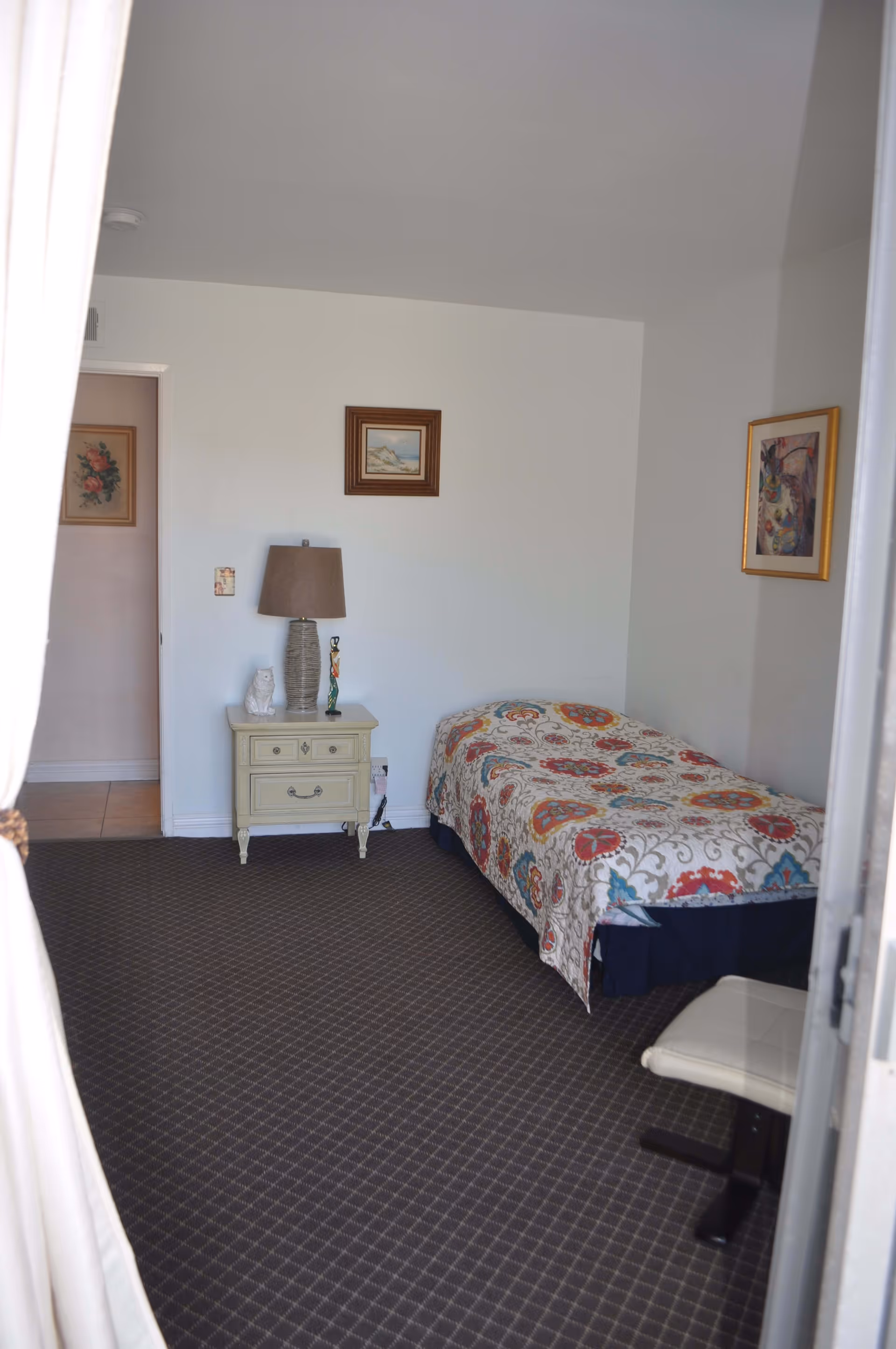 A small bedroom with a single bed covered in a colorful patterned quilt. Next to the bed is a cream-colored nightstand with a lamp, a small owl figurine, and another decorative item. The walls are white and decorated with three framed pictures. The floor is carpeted with a dark patterned carpet. A white chair is partially visible in the foreground.